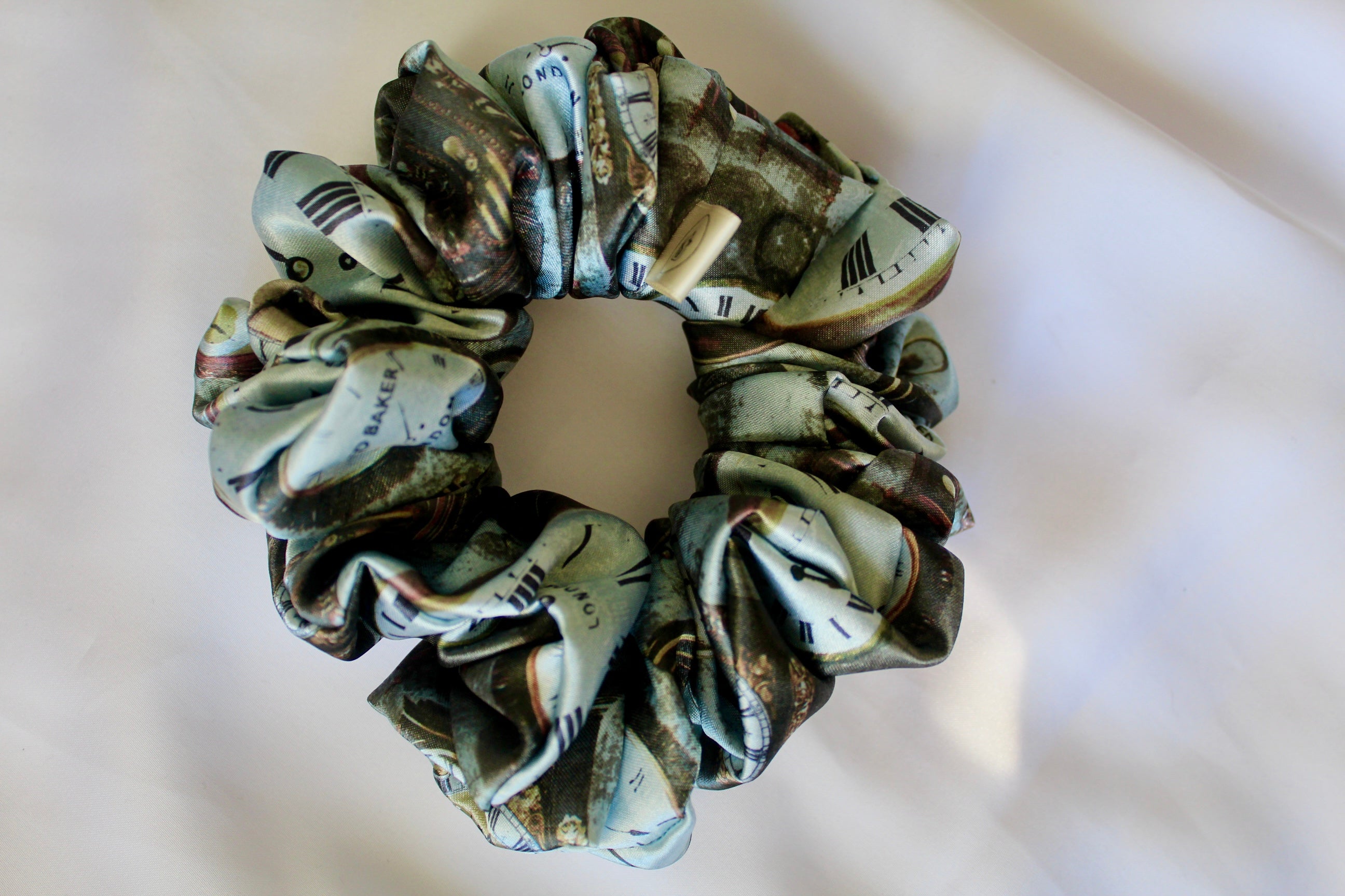 This image features a handmade regular sized hair scrunchie with a vintage clock print on a satin fabric. The scrunchie is designed with a mix of antique- style timepieces, numbers and text elements, giving it a steampunk and Victorian Aesthetic. The primary colours include shades of brown and beige. The fabric has a slight sheen adding a luxurious touch to the accessory. Ideal for those looking for a statement hair accessory.