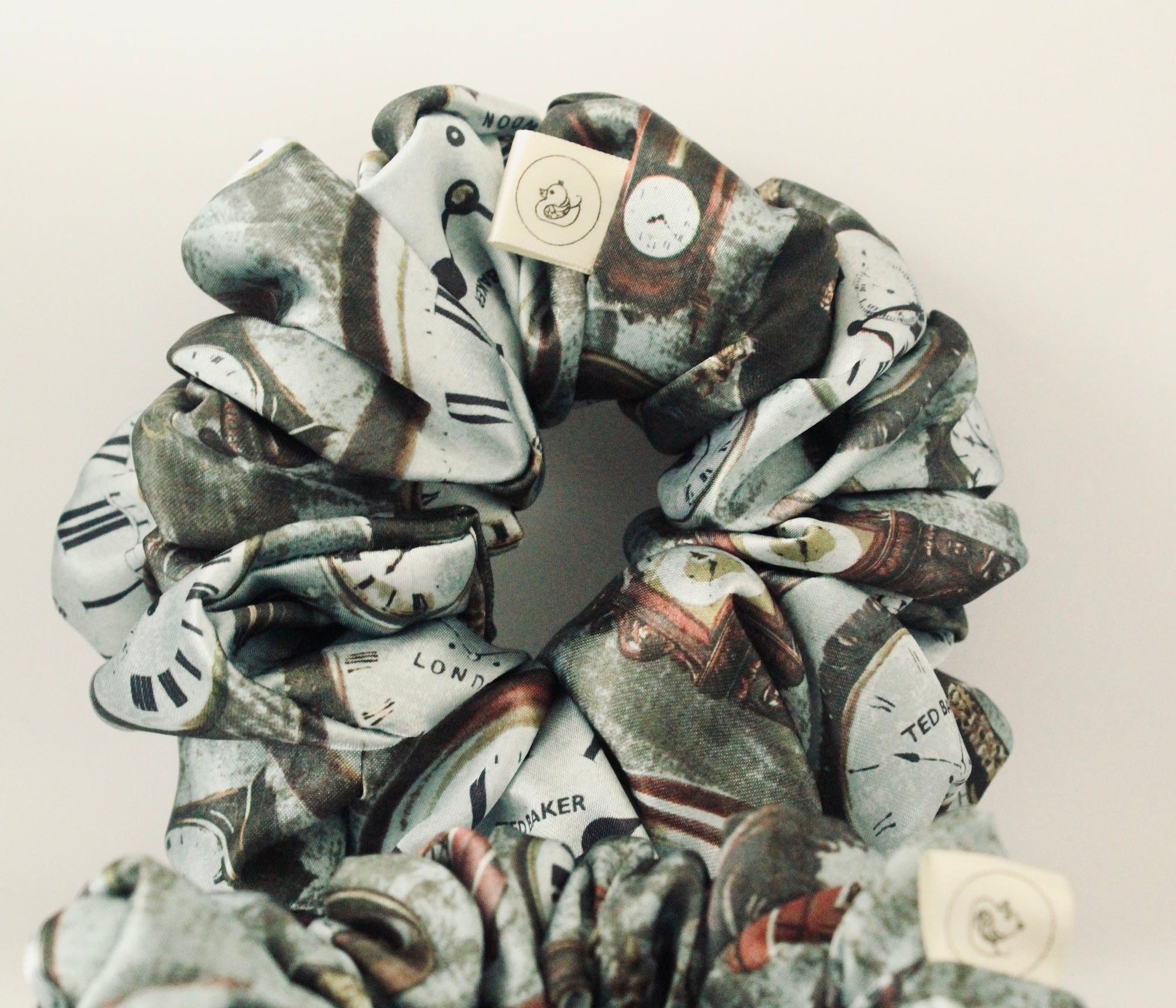 This image features a handmade regular sized hair scrunchie with a vintage clock print on a satin fabric. The scrunchie is designed with a mix of antique- style timepieces, numbers and text elements, giving it a steampunk and Victorian Aesthetic. The primary colours include shades of brown and beige. The fabric has a slight sheen adding a luxurious touch to the accessory. Ideal for those looking for a statement hair accessory.