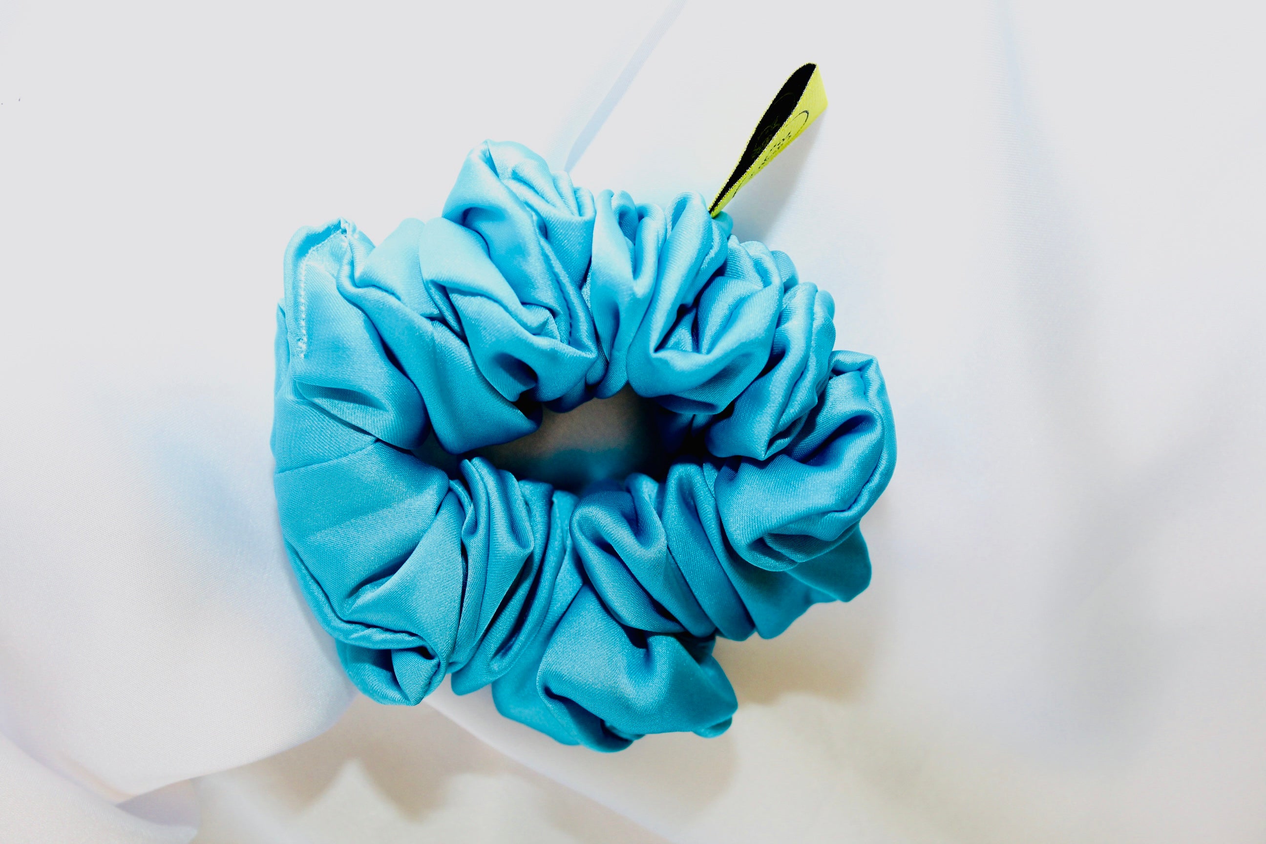 This shows a luxurious scrunchie made of silky textured fabric in a Turquoise Blue giving it an elegant and slightly reflective appearance.The scrunchie is gathered and voluminous creating soft folds and a plush look.A small yellow label tag with a picture of a duck is attached to the scrunchie adding a touch of branding.The overall aesthetic is soft, elegant and stylish. Perfect for those with sensitive scalp or hair loss