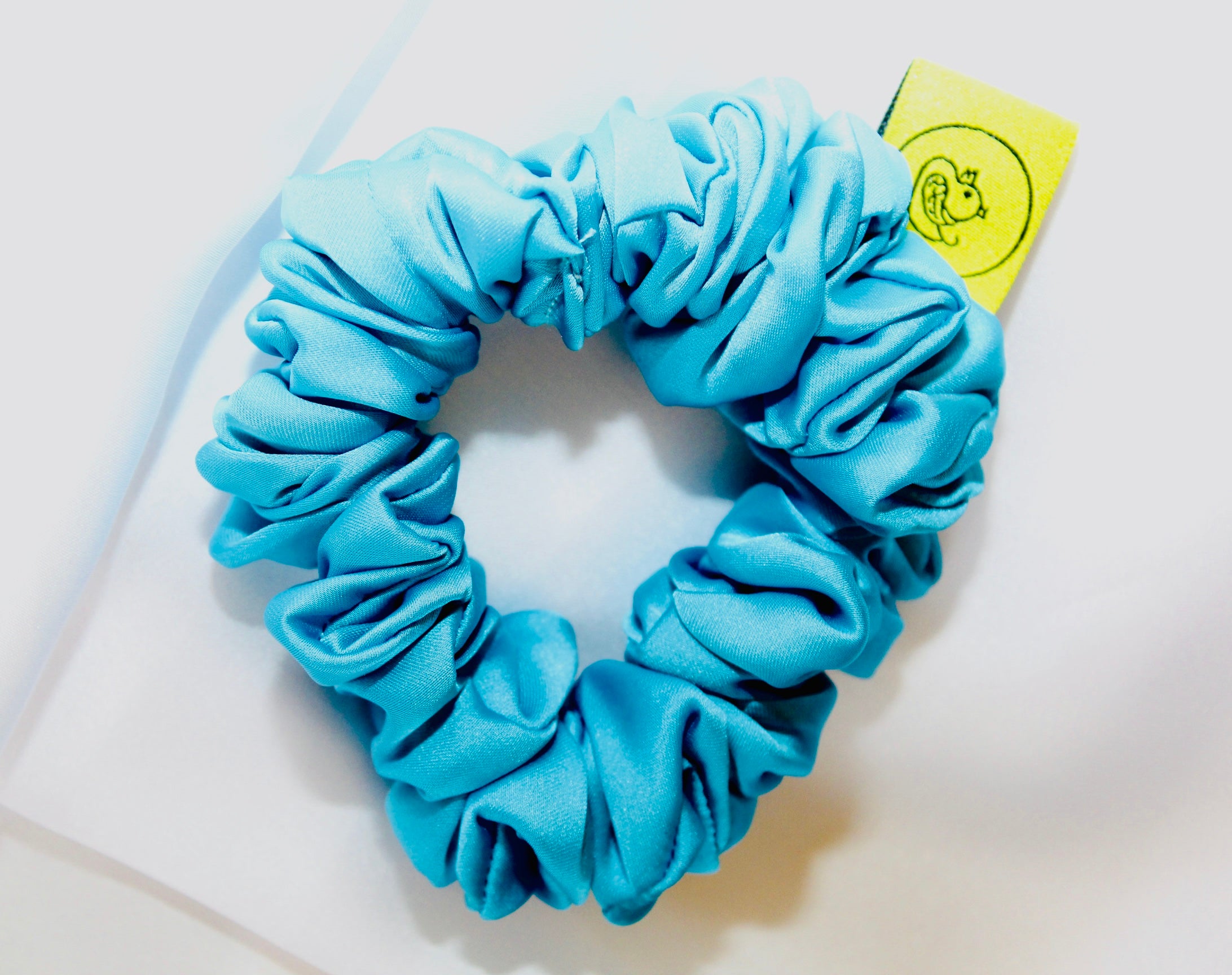 This shows a luxurious scrunchie made of silky textured fabric in a Turquoise Blue giving it an elegant and slightly reflective appearance.The scrunchie is gathered and voluminous creating soft folds and a plush look.A small yellow label tag with a picture of a duck is attached to the scrunchie adding a touch of branding.The overall aesthetic is soft, elegant and stylish. Perfect for those with sensitive scalp or hair loss