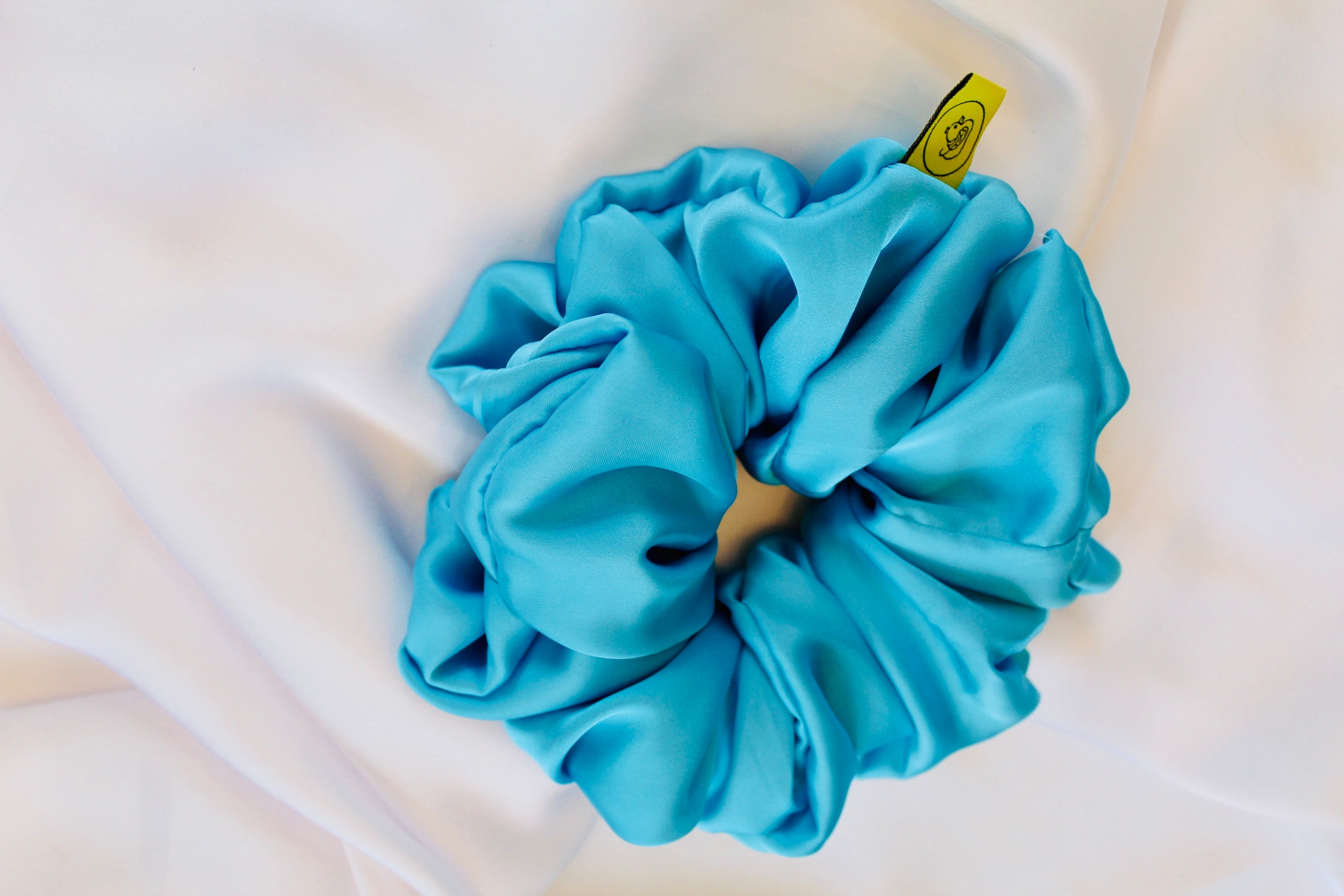 This shows a luxurious scrunchie made of silky textured fabric in a Turquoise Blue giving it an elegant and slightly reflective appearance.The scrunchie is gathered and voluminous creating soft folds and a plush look.A small yellow label tag with a picture of a duck is attached to the scrunchie adding a touch of branding.The overall aesthetic is soft, elegant and stylish. Perfect for those with sensitive scalp or hair loss