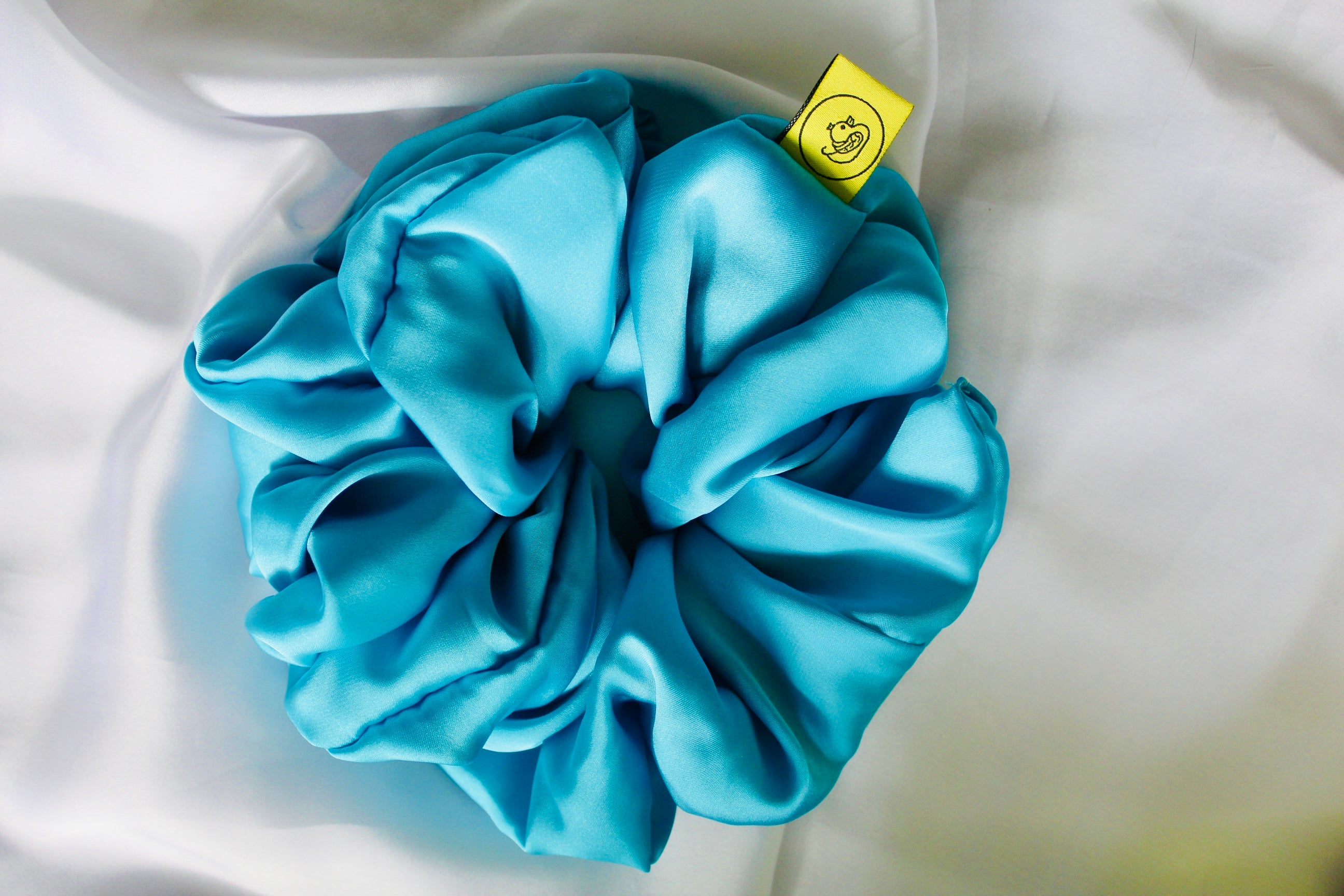 This shows a luxurious scrunchie made of silky textured fabric in a Turquoise Blue giving it an elegant and slightly reflective appearance.The scrunchie is gathered and voluminous creating soft folds and a plush look.A small yellow label tag with a picture of a duck is attached to the scrunchie adding a touch of branding.The overall aesthetic is soft, elegant and stylish. Perfect for those with sensitive scalp or hair loss