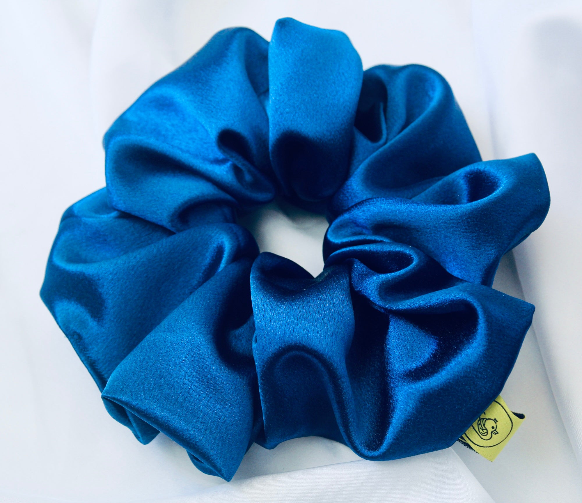 A satin scrunchie in a striking royal blue shade with a silky glossy texture and a ruched design. Crafted from satin this scrunchie feels smooth and cool to the touch. The bold royal blue colour stands out beautifully, making it a statement piece for both casual and evening looks. The gathered satin fabric creates a full textured effect over the elastic ensuring a secure hold.
