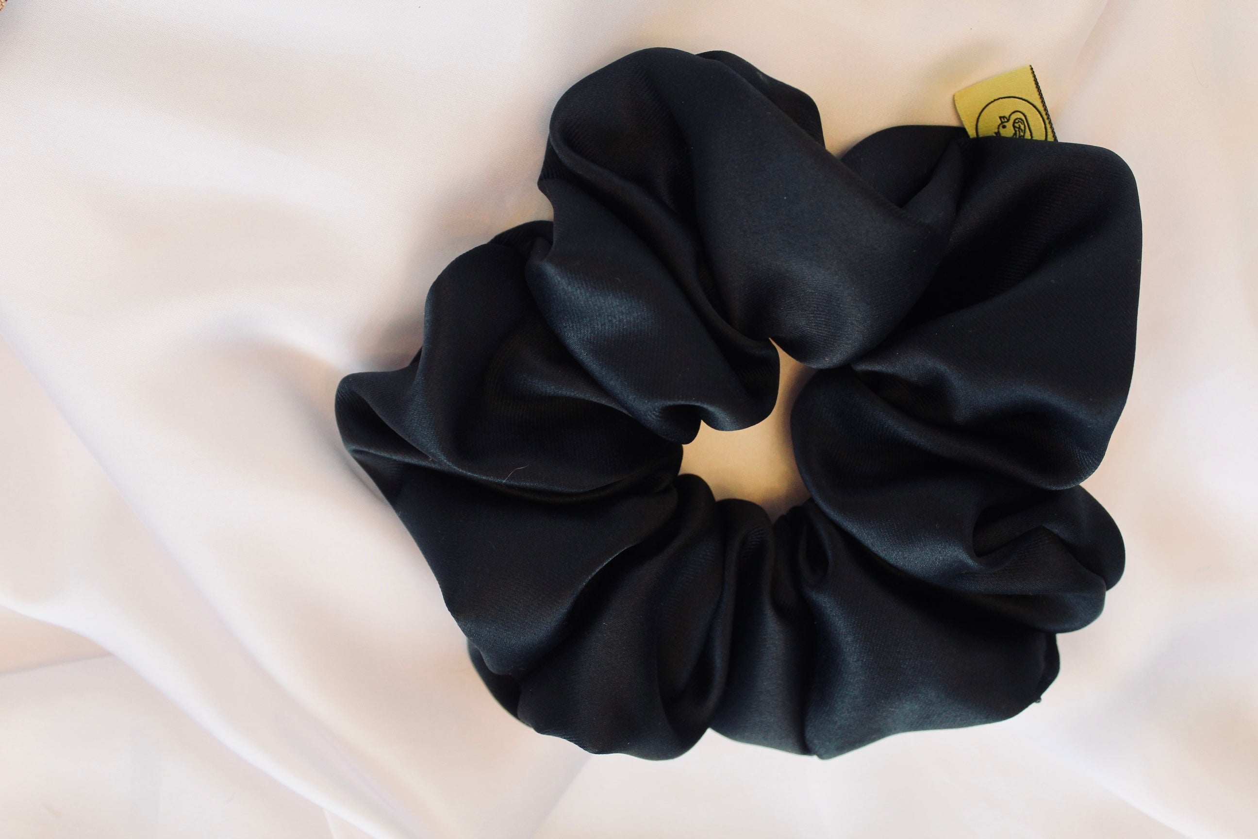 This shows a luxurious scrunchie made of silky textured fabric in a black  thick fabric giving it an elegant and slightly reflective appearance.The scrunchie is gathered and voluminous creating soft folds and a plush look.A small yellow label tag with a picture of a duck is attached to the scrunchie adding a touch of branding.The overall aesthetic is soft, elegant and stylish. Perfect for those with sensitive scalp or hair loss
