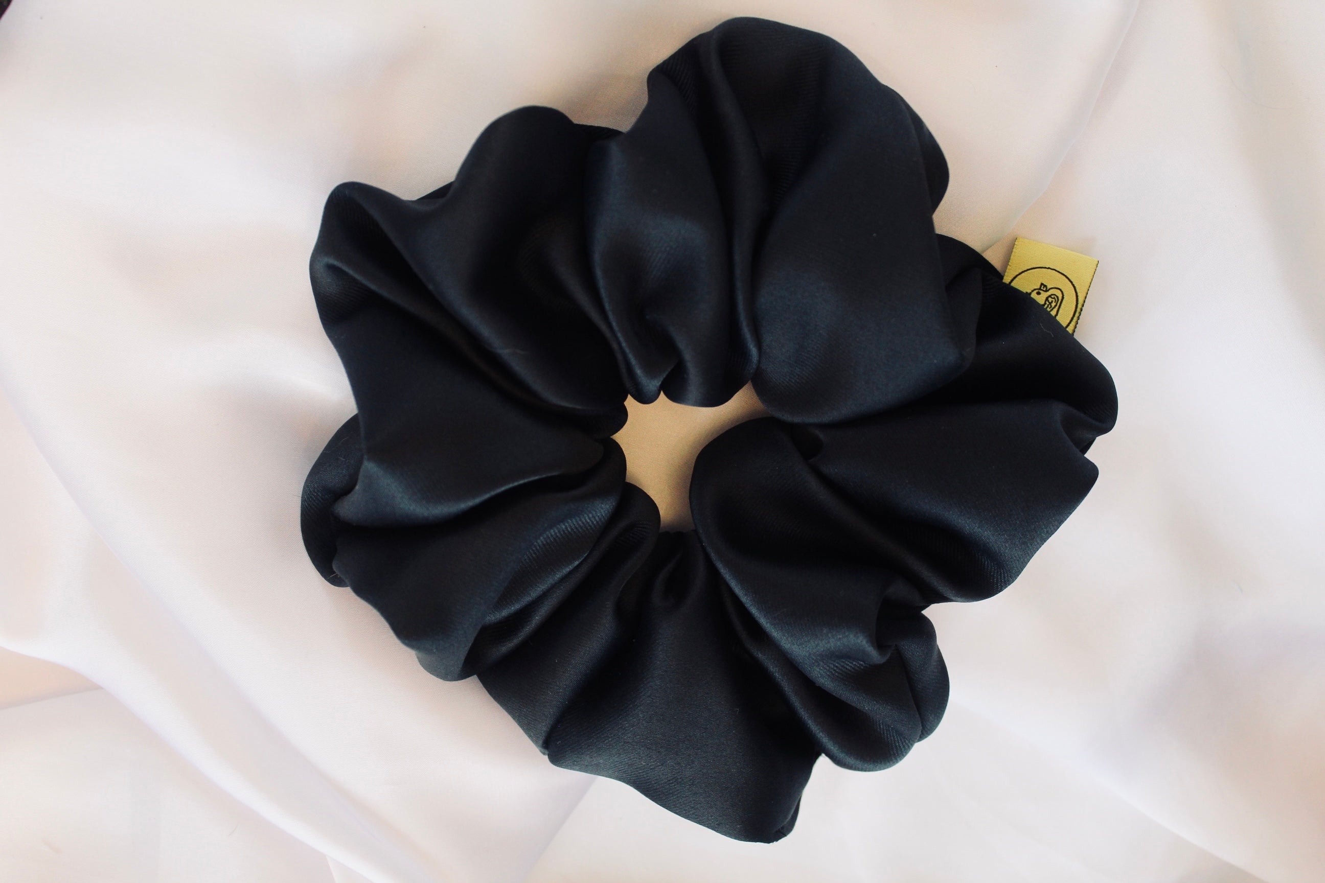 This shows a luxurious scrunchie made of silky textured fabric in a black  thick fabric giving it an elegant and slightly reflective appearance.The scrunchie is gathered and voluminous creating soft folds and a plush look.A small yellow label tag with a picture of a duck is attached to the scrunchie adding a touch of branding.The overall aesthetic is soft, elegant and stylish. Perfect for those with sensitive scalp or hair loss