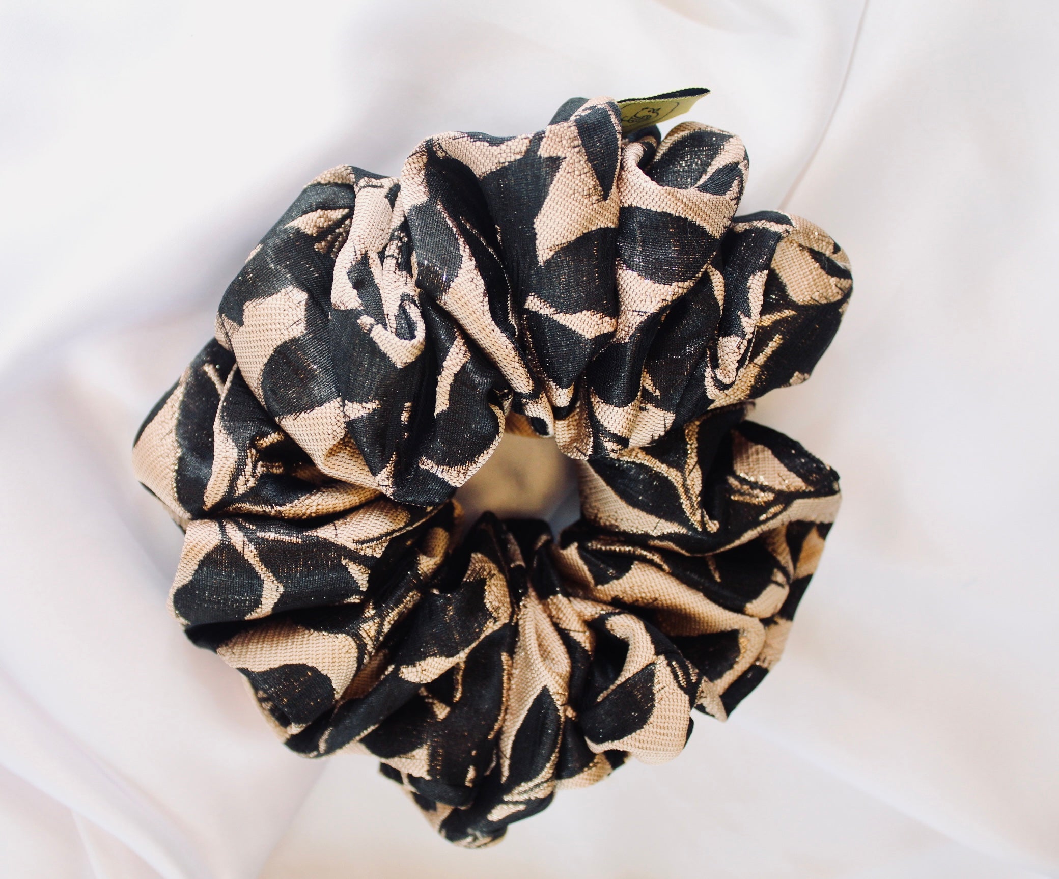 This shows a luxurious scrunchie made of silky textured fabric in a black  and gold woven fabric giving it an elegant and slightly reflective appearance.The scrunchie is gathered and voluminous creating soft folds and a plush look.A small yellow label tag with a picture of a duck is attached to the scrunchie adding a touch of branding.The overall aesthetic is soft, elegant and stylish. Perfect for those with sensitive scalp or hair loss