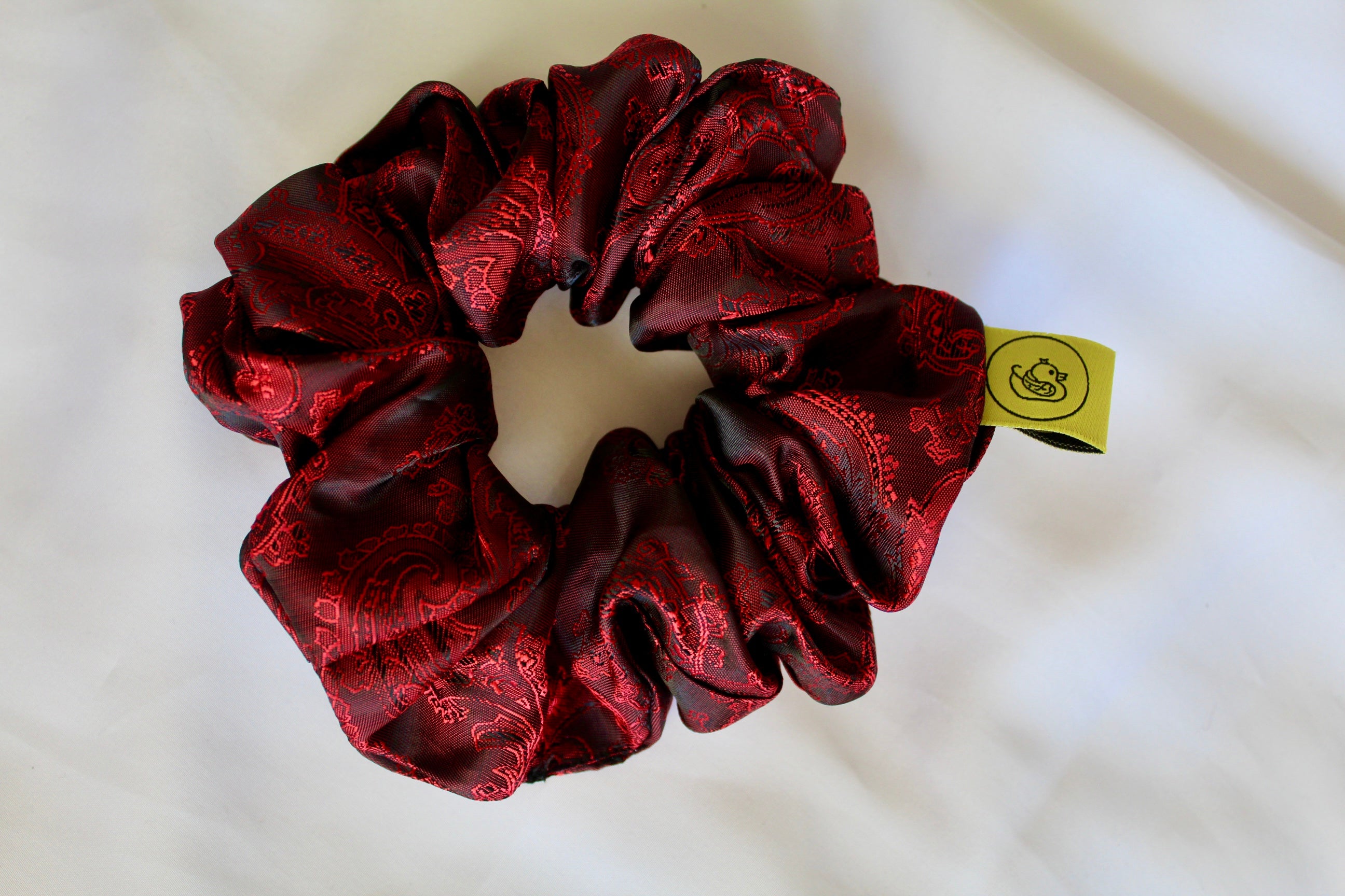 This shows a luxurious scrunchie made of silky textured fabric in a red and black colour.The fabric has a subtle paisley pattern wove into it, giving it an elegant and slightly reflective appearance.The scrunchie is gathered and voluminous creating soft folds and a plush look.A small yellow label tag with a picture of a duck is attached to the scrunchie adding a touch of branding.The overall aesthetic is soft, elegant and stylish. Perfect for those with sensitive scalp or hair loss