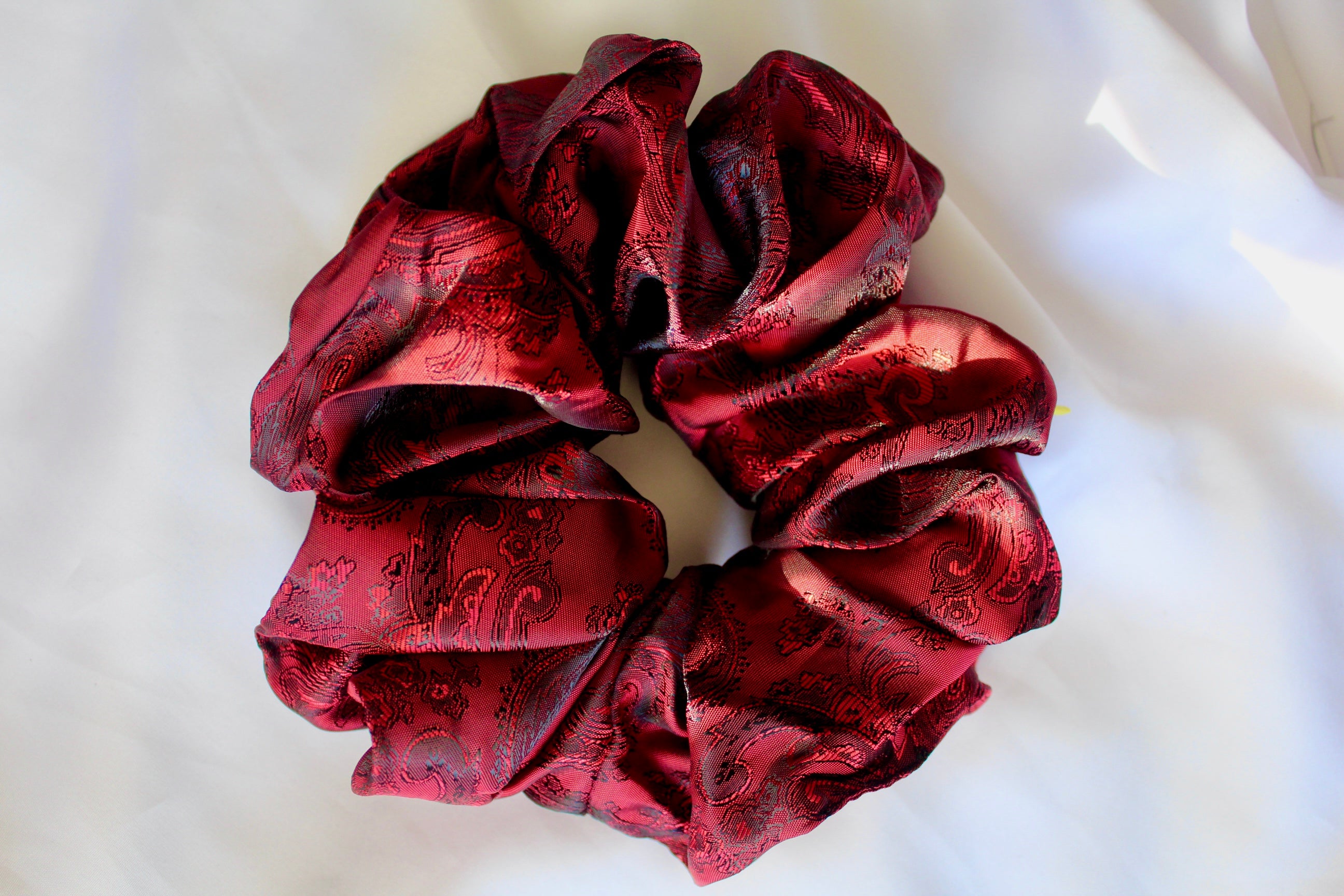 This shows a luxurious scrunchie made of silky textured fabric in a red and black colour.The fabric has a subtle paisley pattern wove into it, giving it an elegant and slightly reflective appearance.The scrunchie is gathered and voluminous creating soft folds and a plush look.A small yellow label tag with a picture of a duck is attached to the scrunchie adding a touch of branding.The overall aesthetic is soft, elegant and stylish. Perfect for those with sensitive scalp or hair loss