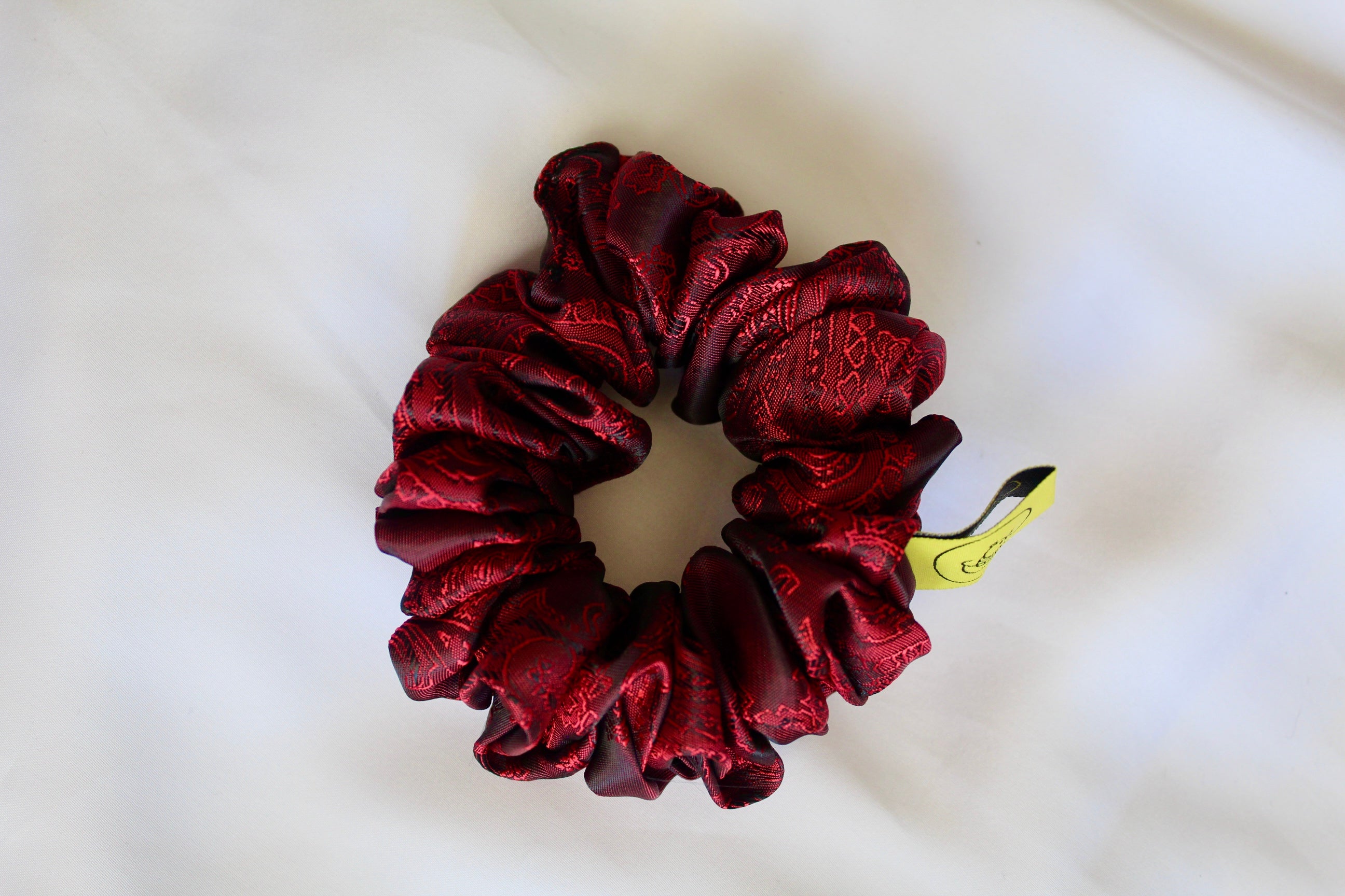 This shows a luxurious scrunchie made of silky textured fabric in a red and black colour.The fabric has a subtle paisley pattern wove into it, giving it an elegant and slightly reflective appearance.The scrunchie is gathered and voluminous creating soft folds and a plush look.A small yellow label tag with a picture of a duck is attached to the scrunchie adding a touch of branding.The overall aesthetic is soft, elegant and stylish. Perfect for those with sensitive scalp or hair loss