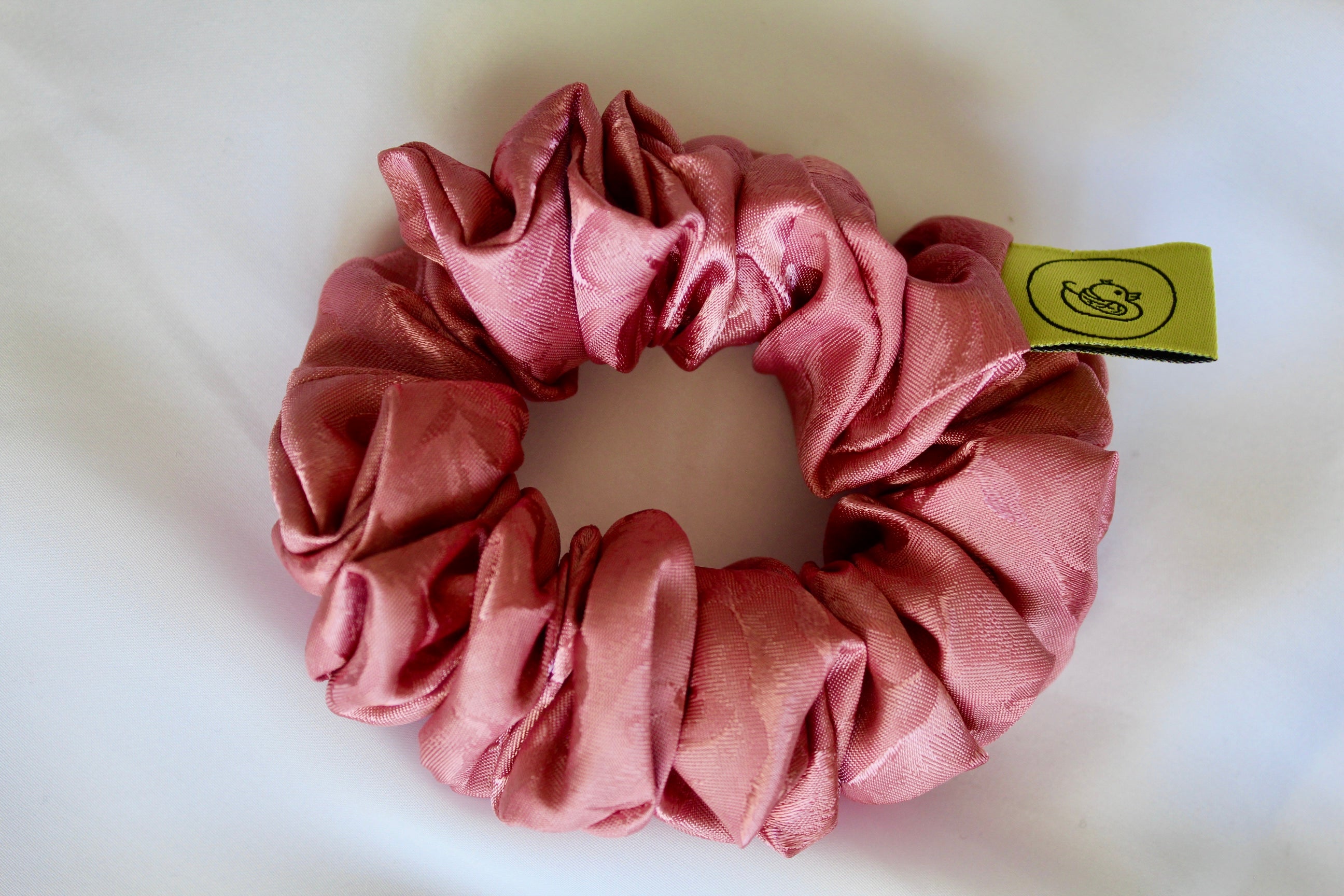 This shows a luxurious scrunchie made of silky textured fabric in a dusty rose pink colour.The fabric has a subtle floral pattern wove into it, giving it an elegant and slightly reflective appearance.The scrunchie is gathered and voluminous creating soft folds and a plush look.A small yellow label tag with a picture of a duck is attached to the scrunchie adding a touch of branding.The overall aesthetic is soft, elegant and stylish.These hair scrunchies are perfect for those with sensitive scalp or hair loss