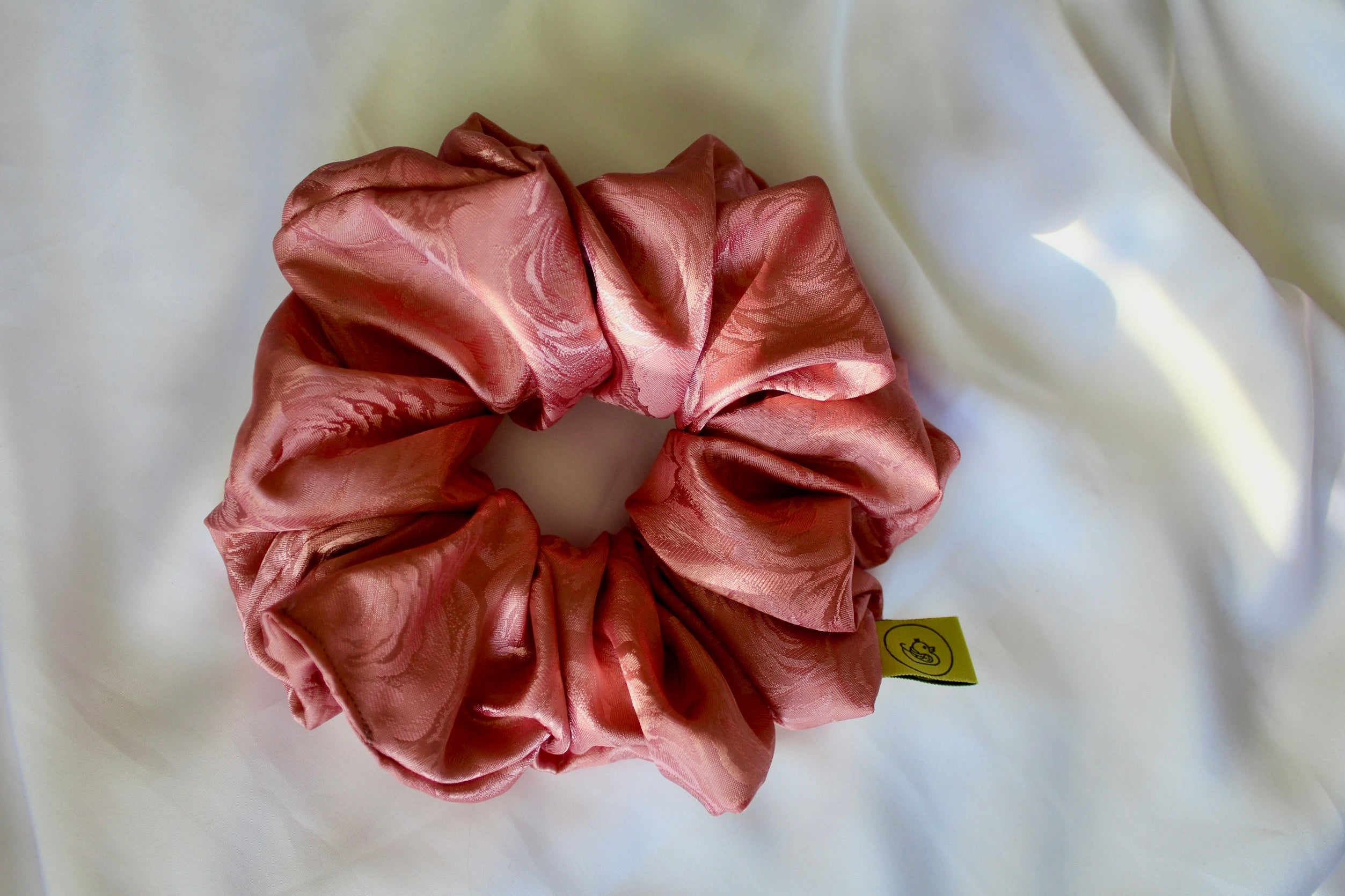 This shows a luxurious scrunchie made of silky textured fabric in a dusty rose pink colour.The fabric has a subtle floral pattern wove into it, giving it an elegant and slightly reflective appearance.The scrunchie is gathered and voluminous creating soft folds and a plush look.A small yellow label tag with a picture of a duck is attached to the scrunchie adding a touch of branding.The overall aesthetic is soft, elegant and stylish.These hair scrunchies are perfect for those with sensitive scalp or hair loss