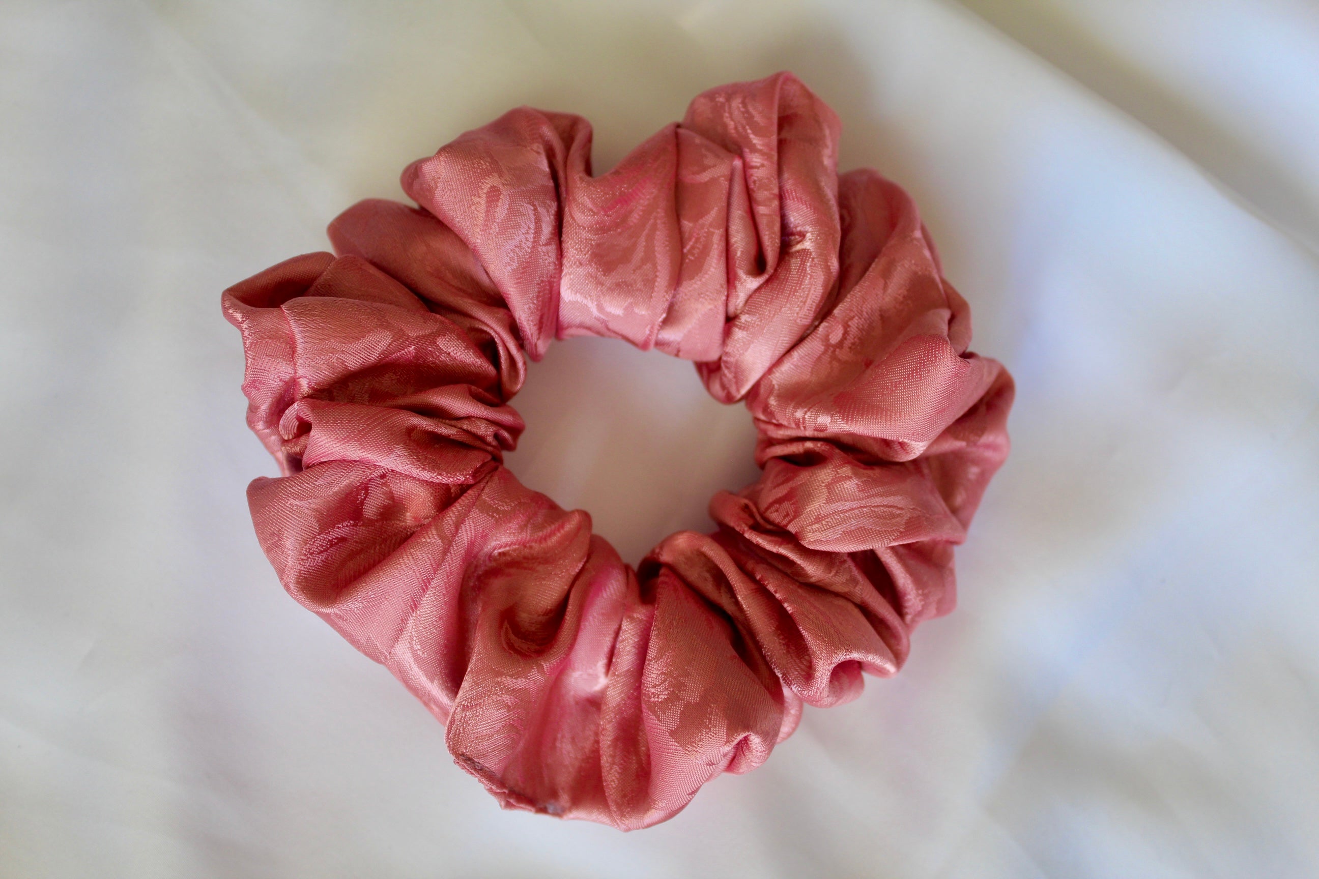 This shows a luxurious scrunchie made of silky textured fabric in a dusty rose pink colour.The fabric has a subtle floral pattern wove into it, giving it an elegant and slightly reflective appearance.The scrunchie is gathered and voluminous creating soft folds and a plush look.A small yellow label tag with a picture of a duck is attached to the scrunchie adding a touch of branding.The overall aesthetic is soft, elegant and stylish.These hair scrunchies are perfect for those with sensitive scalp or hair loss