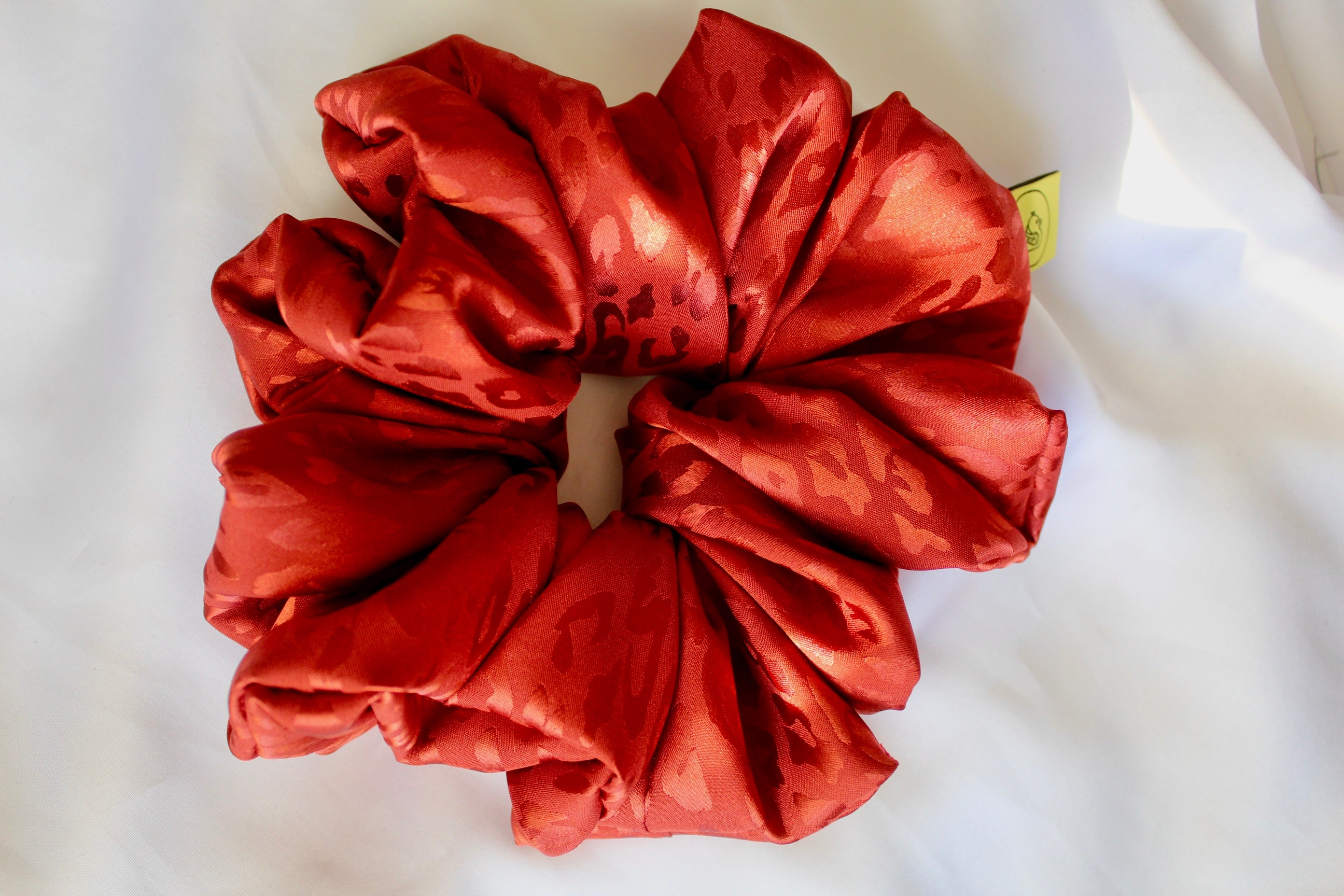 This shows a luxurious scrunchie made of silky textured fabric in a burnt orange/red colour.The fabric has a subtle leopard print pattern wove into it, giving it an elegant and slightly reflective appearance.The scrunchie is gathered and voluminous creating soft folds and a plush look.A small yellow label tag with a picture of a duck is attached to the scrunchie adding a touch of branding.The overall aesthetic is soft, elegant and stylish. Perfect for those with sensitive scalp or hair loss