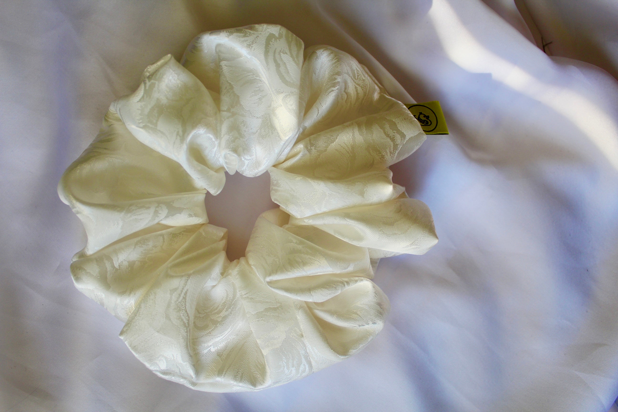 This shows a luxurious scrunchie made of silky textured fabric in a cream ivory colour.The fabric has a subtle floral pattern wove into it, giving it an elegant and slightly reflective appearance.The scrunchie is gathered and voluminous creating soft folds and a plush look.A small yellow label tag with a picture of a duck is attached to the scrunchie adding a touch of branding.The overall aesthetic is soft, elegant and stylish.These hair scrunchies are perfect for those with sensitive scalp or hair loss