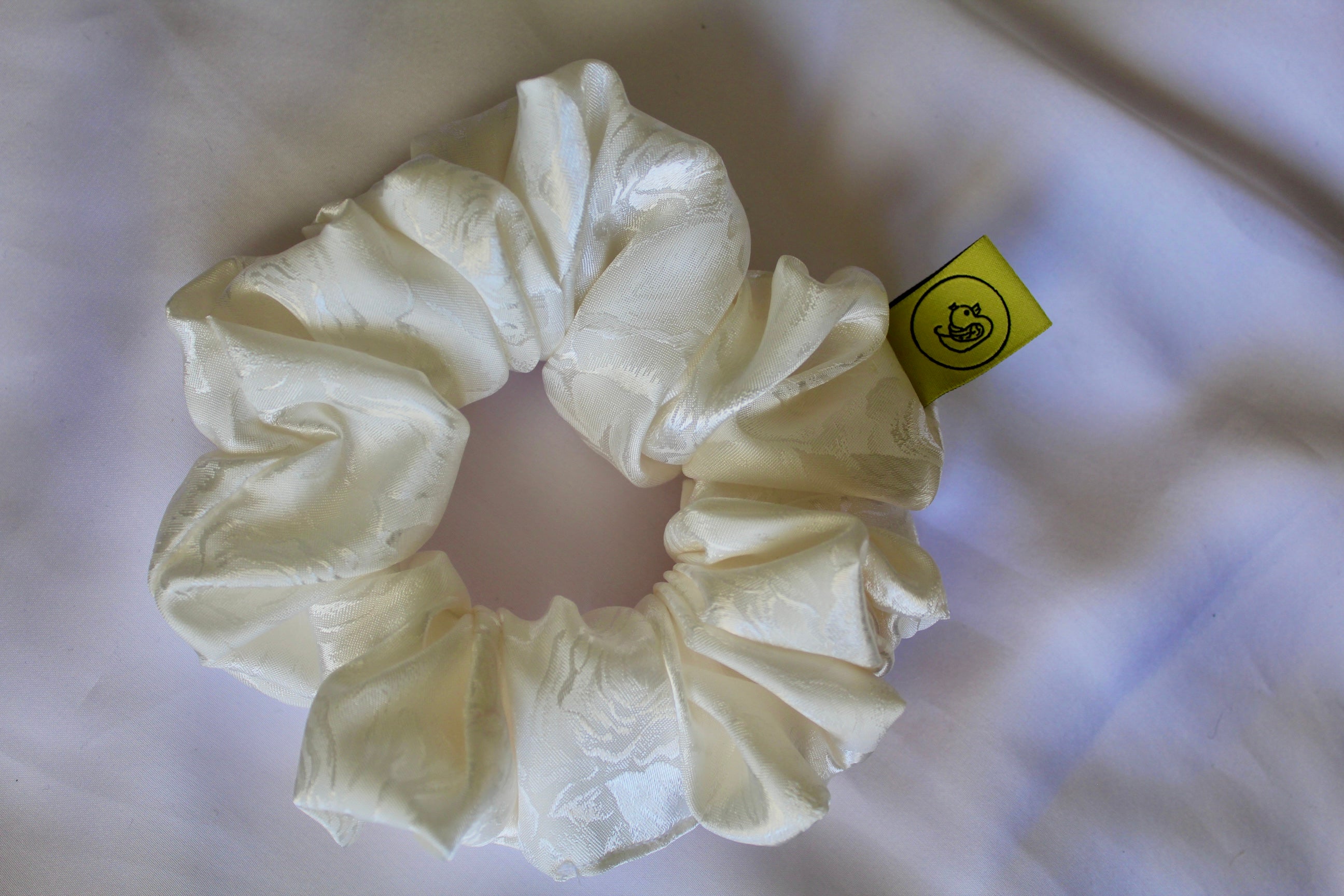 This shows a luxurious scrunchie made of silky textured fabric in a cream ivory colour.The fabric has a subtle floral pattern wove into it, giving it an elegant and slightly reflective appearance.The scrunchie is gathered and voluminous creating soft folds and a plush look.A small yellow label tag with a picture of a duck is attached to the scrunchie adding a touch of branding.The overall aesthetic is soft, elegant and stylish.These hair scrunchies are perfect for those with sensitive scalp or hair loss
