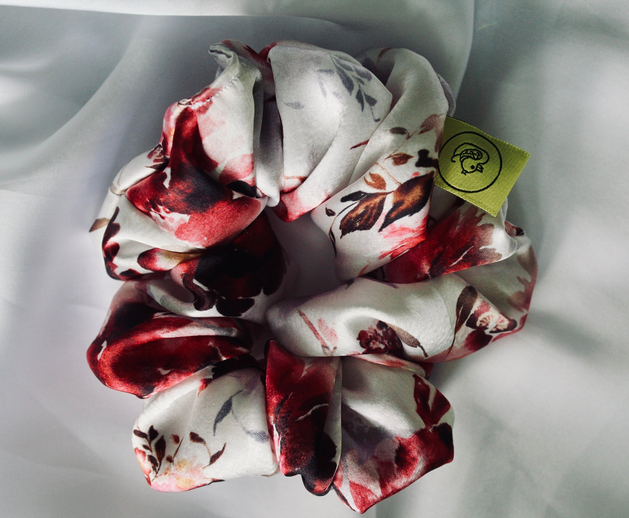 This shows a luxurious scrunchie made of silky textured fabric in a dusty pink and red floral pattern. The fabric has a subtle floral pattern wove into it, giving it an elegant and slightly reflective appearance.The scrunchie is gathered and voluminous creating soft folds and a plush look.A small yellow label tag with a picture of a duck is attached to the scrunchie adding a touch of branding.The overall aesthetic is soft, elegant and stylish. Perfect for those with sensitive scalp or hair loss