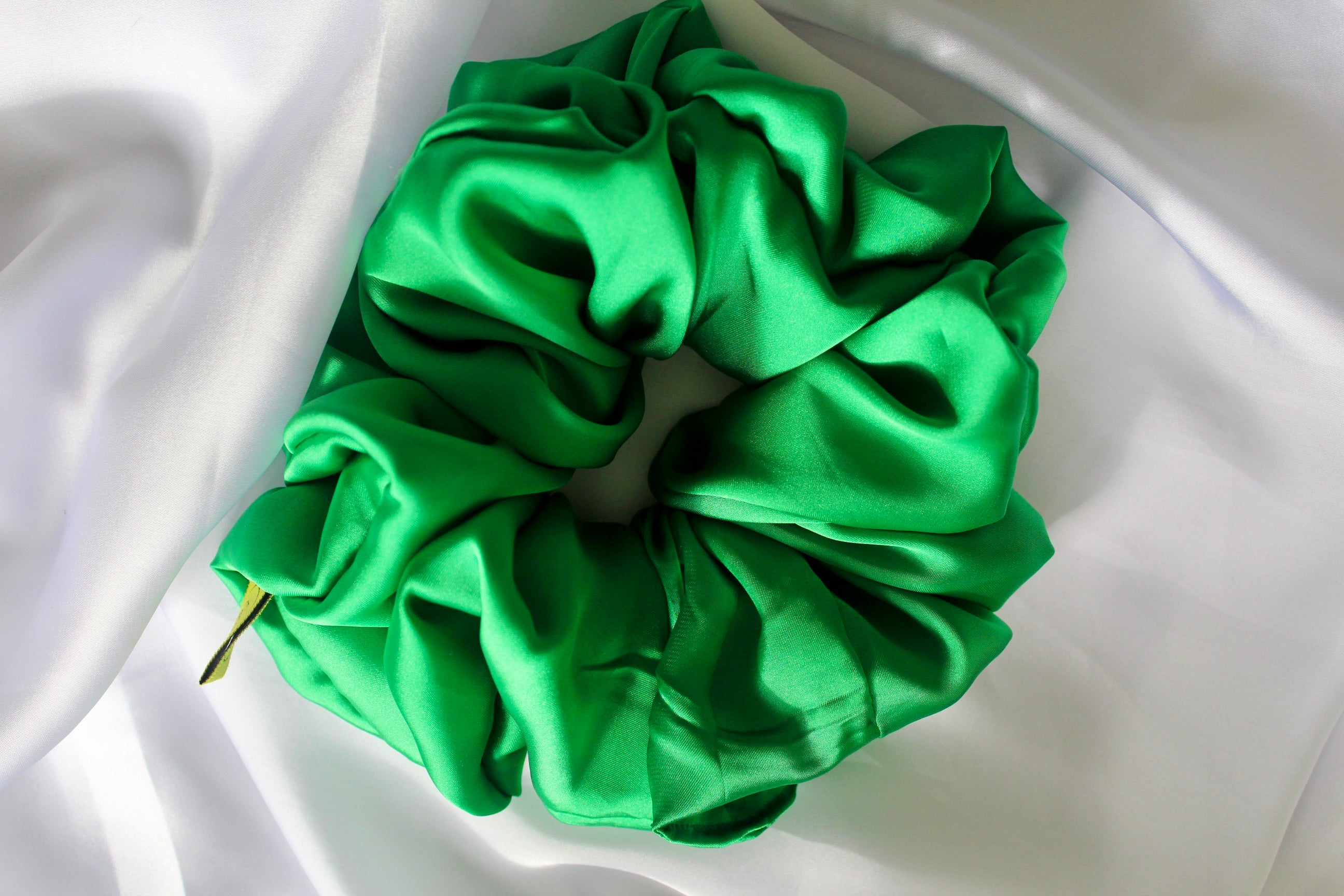 This shows a luxurious scrunchie made of silky textured fabric in a Emerald Green giving it an elegant and slightly reflective appearance.The scrunchie is gathered and voluminous creating soft folds and a plush look.A small yellow label tag with a picture of a duck is attached to the scrunchie adding a touch of branding.The overall aesthetic is soft, elegant and stylish. Perfect for those with sensitive scalp or hair loss