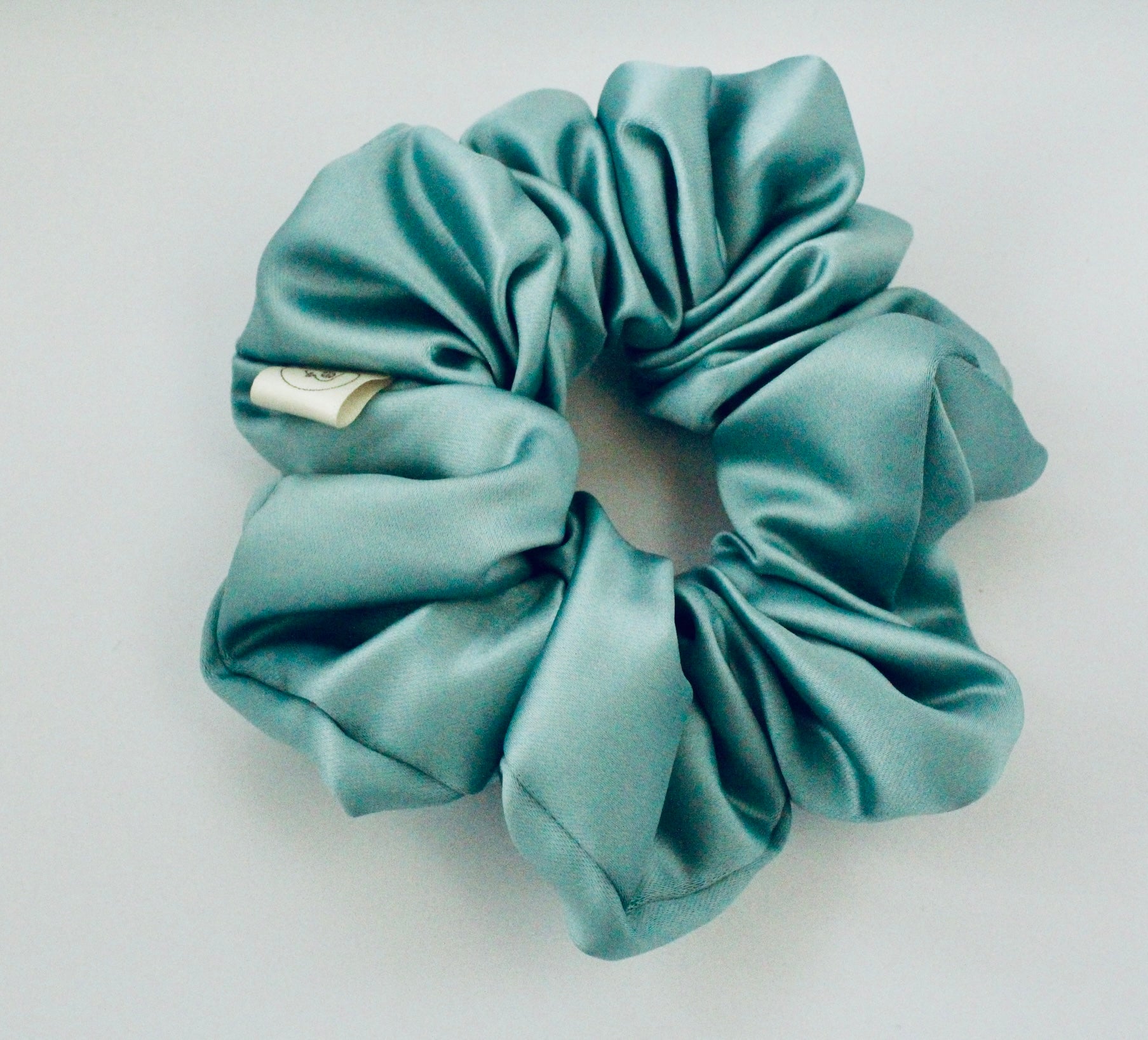 This shows a luxurious scrunchie made of silky textured fabric in an mint green running through the fabric giving it an elegant and slightly reflective appearance.The scrunchie is gathered and voluminous creating soft folds and a plush look.A small yellow label tag with a picture of a duck is attached to the scrunchie adding a touch of branding.The overall aesthetic is soft, elegant and stylish. Perfect for those with sensitive scalp or hair loss