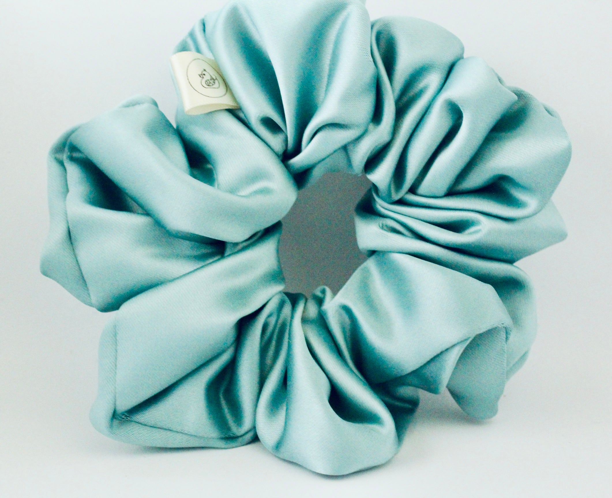 This shows a luxurious scrunchie made of silky textured fabric in an mint green running through the fabric giving it an elegant and slightly reflective appearance.The scrunchie is gathered and voluminous creating soft folds and a plush look.A small yellow label tag with a picture of a duck is attached to the scrunchie adding a touch of branding.The overall aesthetic is soft, elegant and stylish. Perfect for those with sensitive scalp or hair loss