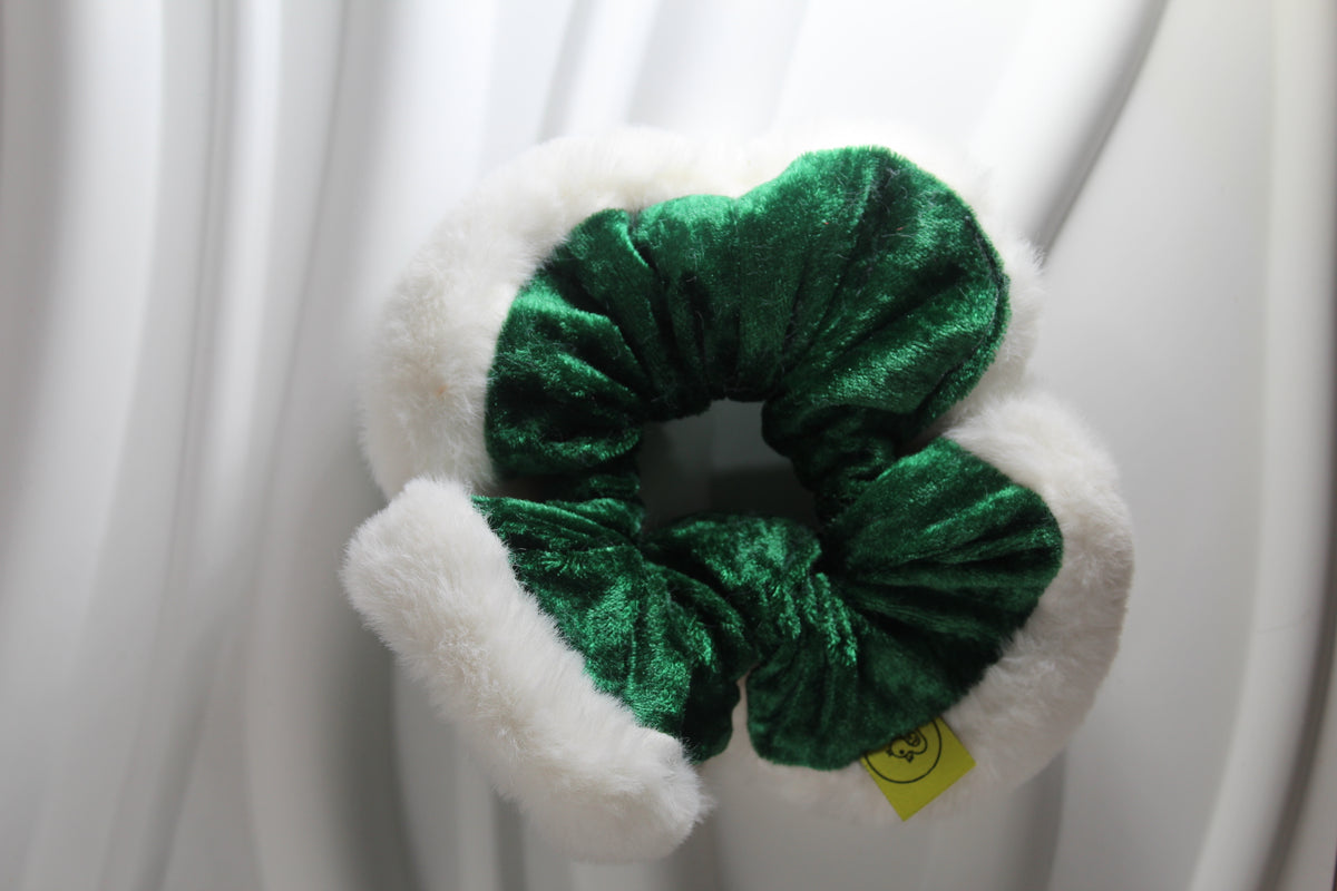 A green velvet scrunchie with white faux fur trim, shown against a draped white fabric background.