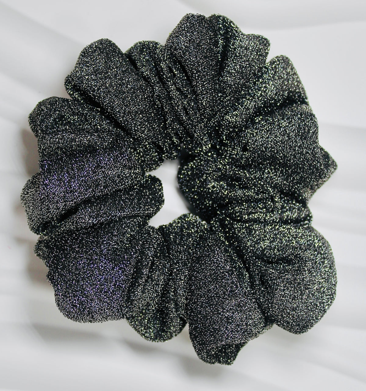 A single black glittery scrunchie with a textured, sparkly finish, photographed flat against a white fabric background.