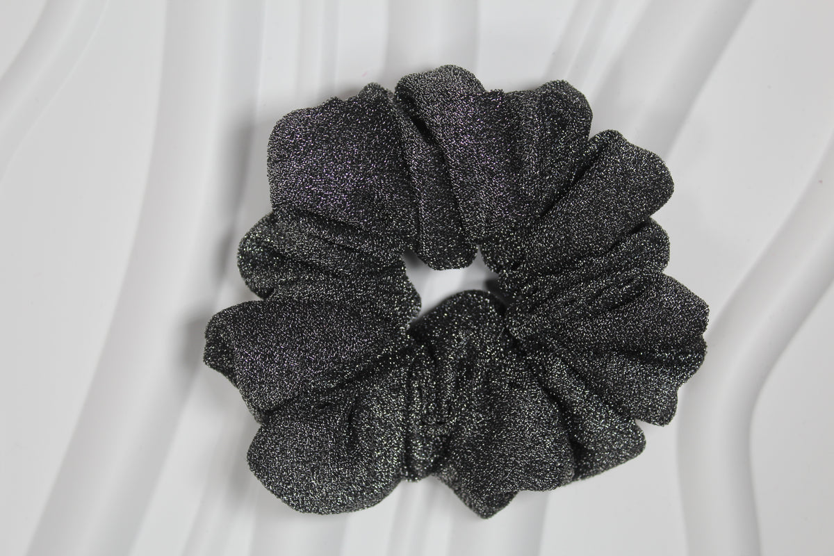 A single black glittery scrunchie with a textured, sparkly finish, photographed from above on a white background with soft draped folds.