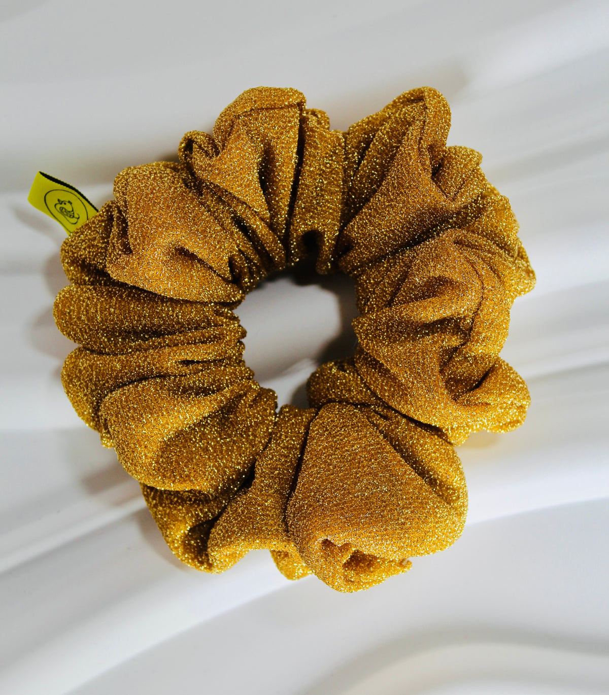 A golden yellow glittery scrunchie with a textured, sparkly fabric, shown on a draped white fabric background with a small yellow tag attached.