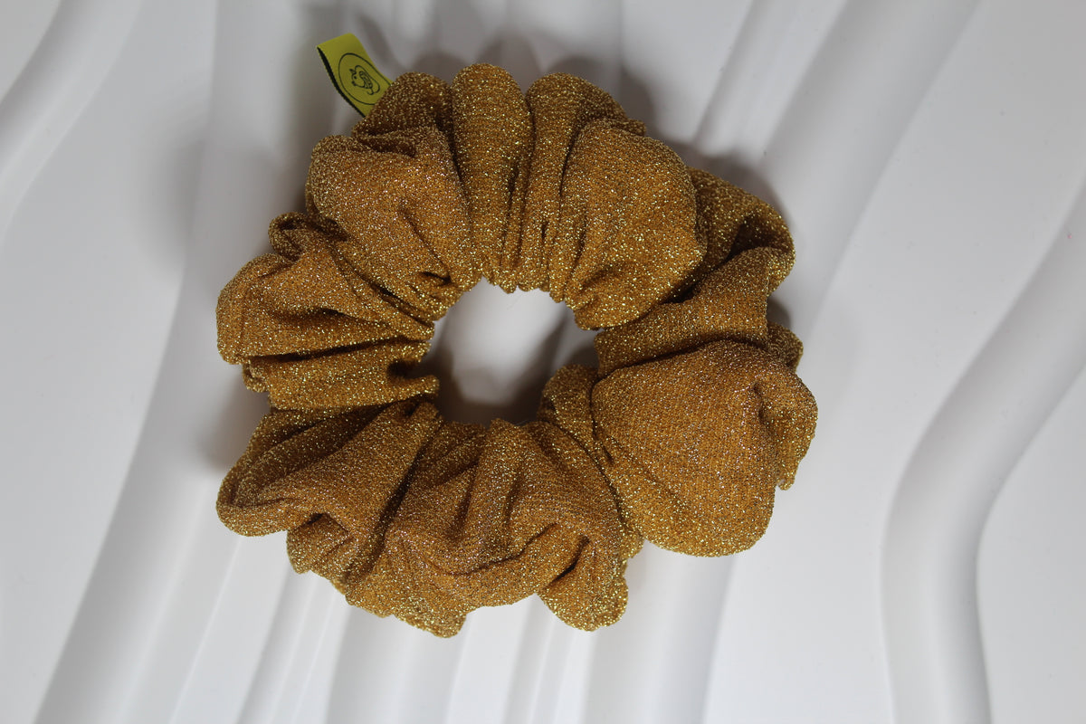 A single metallic gold scrunchie with a glittery texture, photographed from above on a white wavy surface.