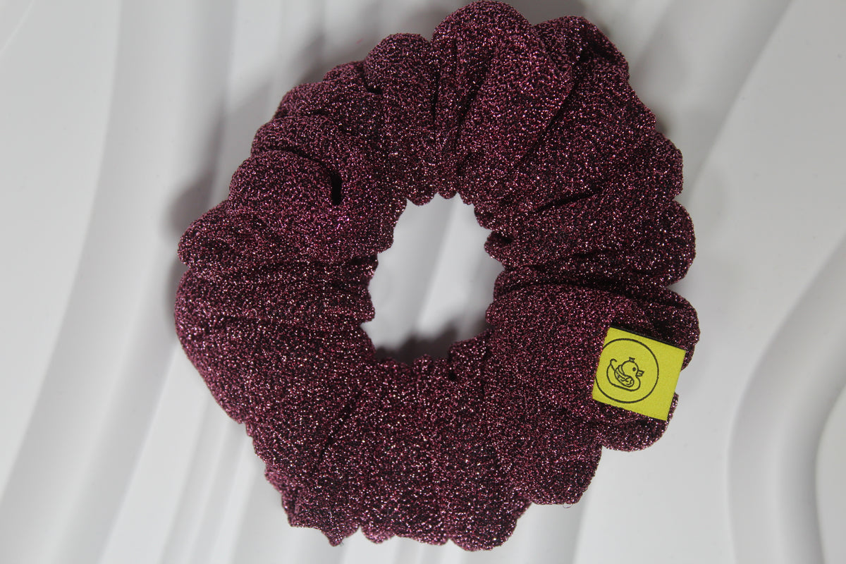 A glittery deep burgundy scrunchie with a small yellow brand tag, photographed on a white wavy background.