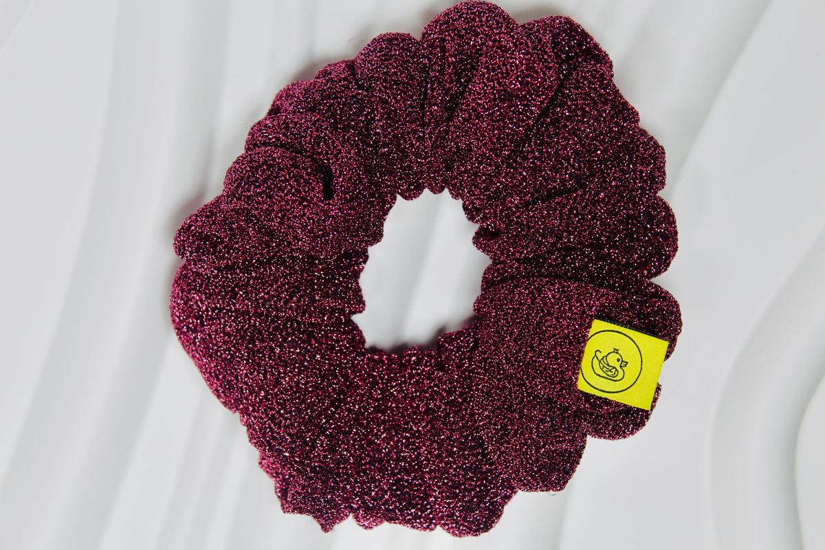 A textured, sparkly deep magenta scrunchie with a small yellow brand tag, photographed on a white wavy fabric background.