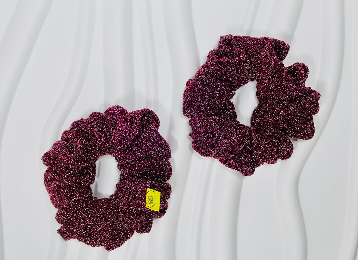 Two textured, glittery deep purple scrunchies on a white wavy background, with one featuring a small yellow tag.