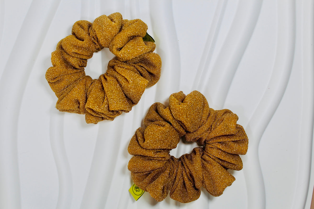 Two glittery mustard-gold scrunchies displayed on a white wavy fabric background.