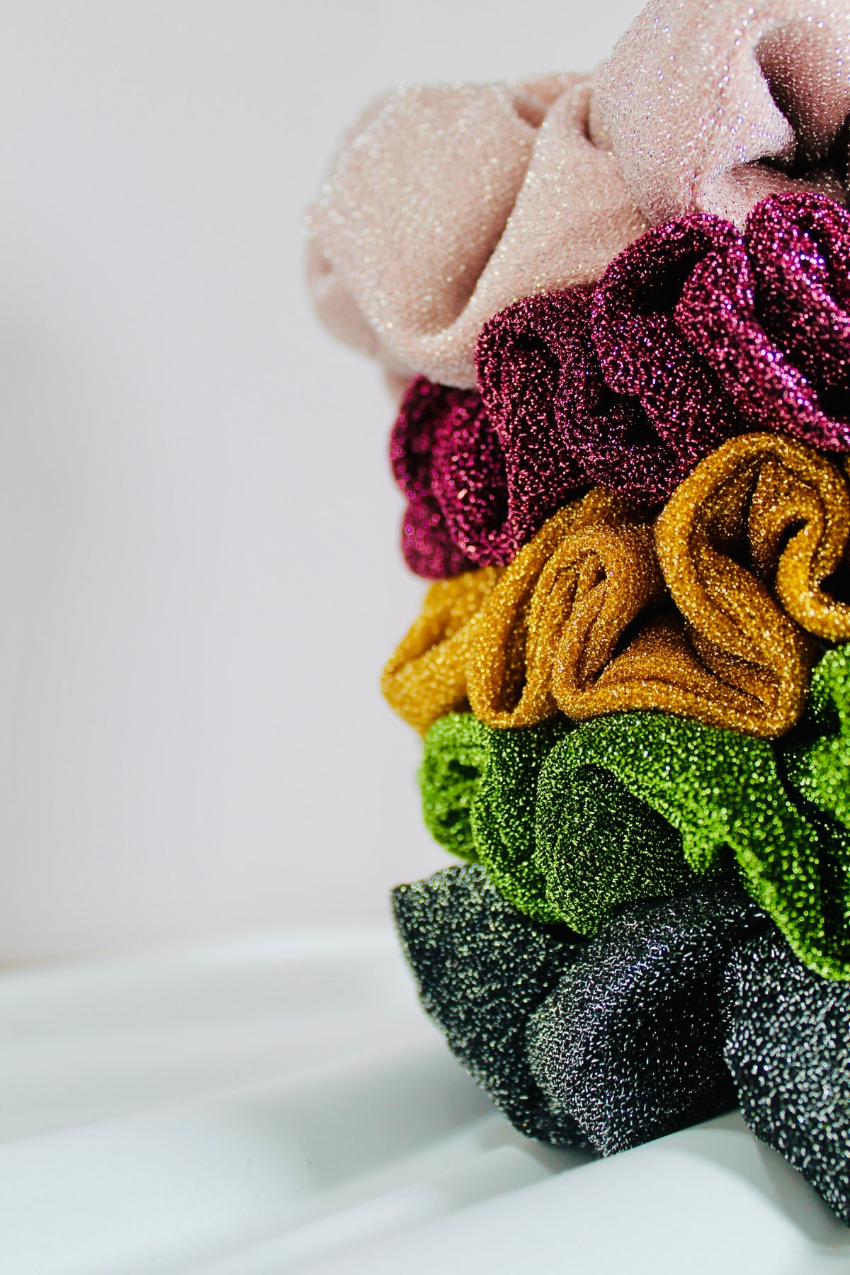 A stack of glittery fabric hair scrunchies in pastel and bright colors, including pink, magenta, mustard yellow, green, and dark gray, photographed against a clean white background.