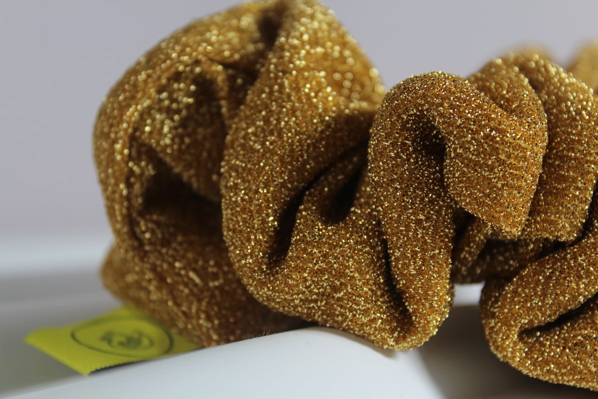 A close-up of a gold glittery scrunchie resting on a white surface with a small yellow tag visible in the lower left corner.