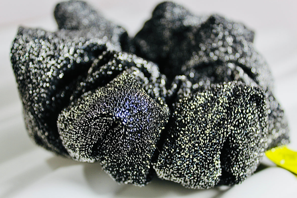 A close-up of a black scrunchie made from highly textured, sparkly metallic fabric that reflects silver and blue light, resting on a white surface.