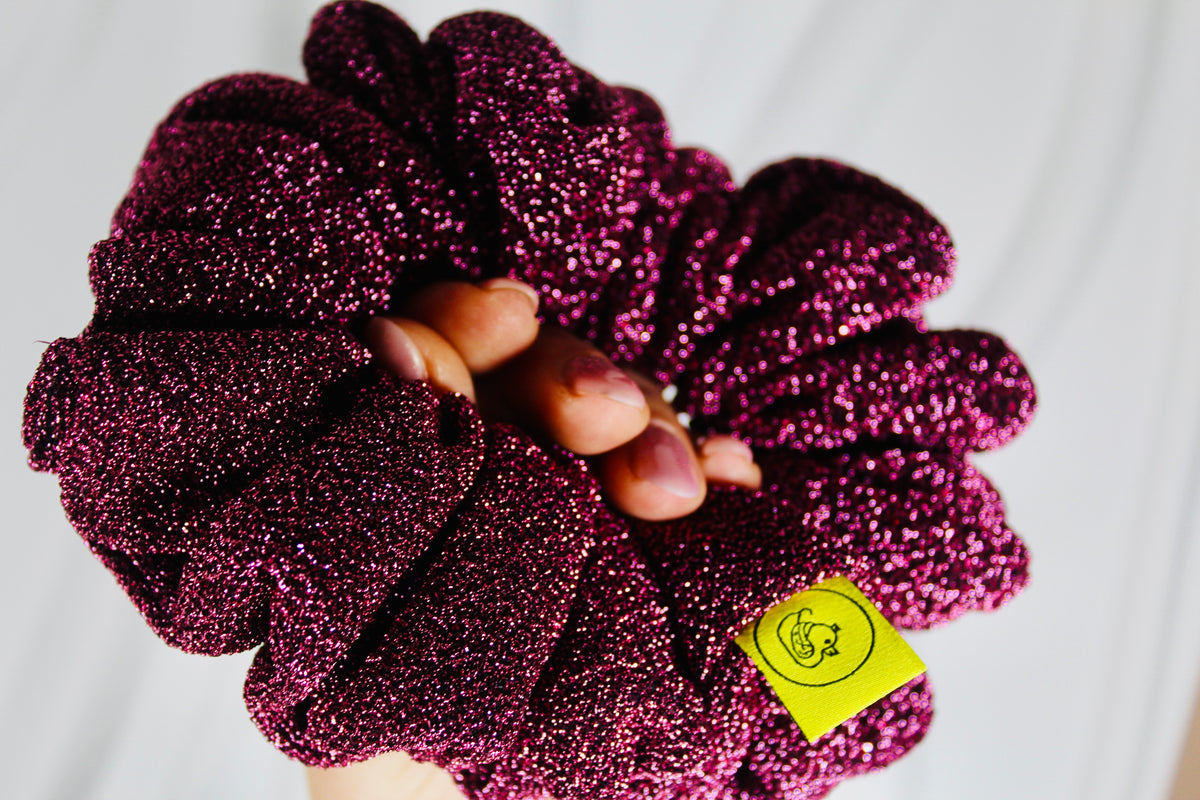 A close-up of a hand holding a large, glittery magenta scrunchie with a small yellow brand tag. The background is a clean, slightly out-of-focus white fabric.