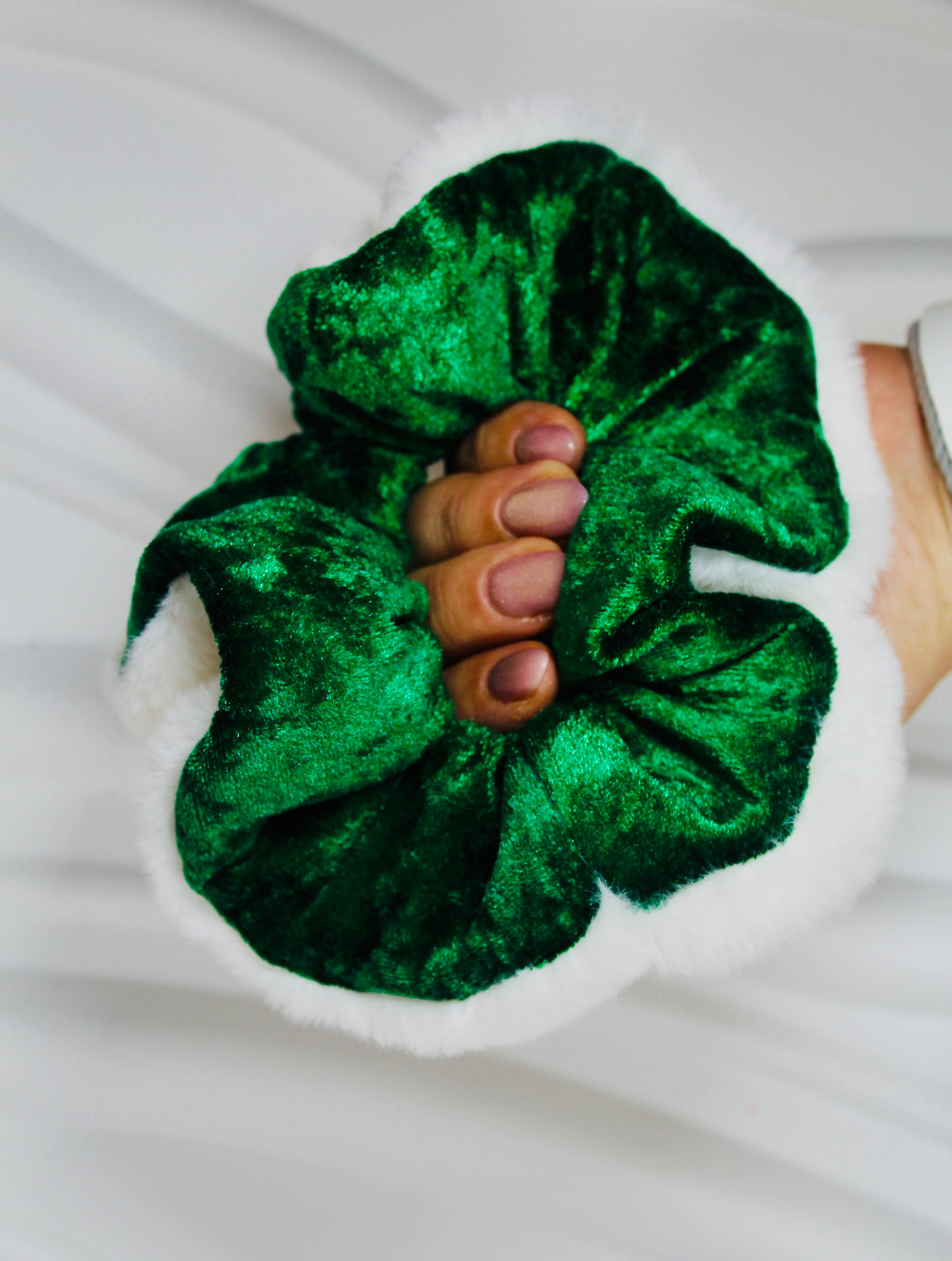 A close-up of a hand holding a green velvet scrunchie with a white faux-fur trim, set against a soft white fabric background.