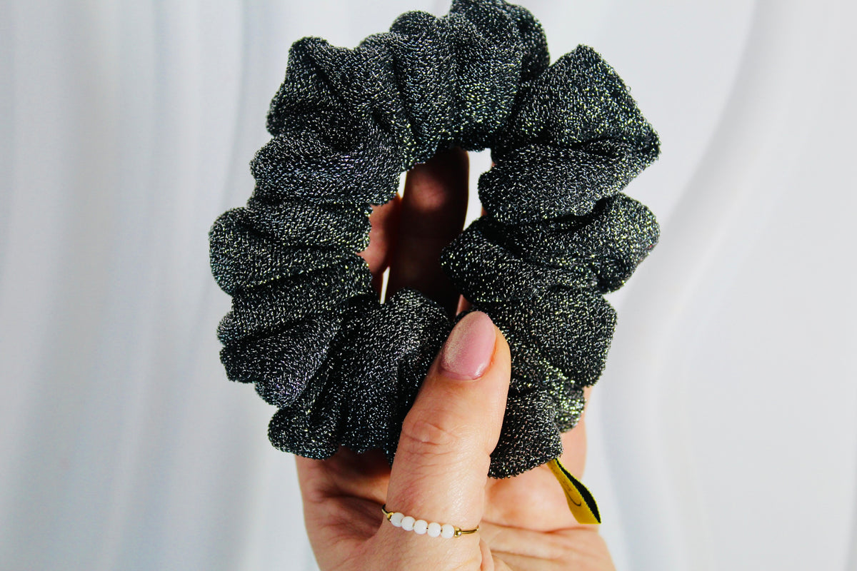 A close-up of a hand holding a dark charcoal gray glittery scrunchie against a soft white draped background.