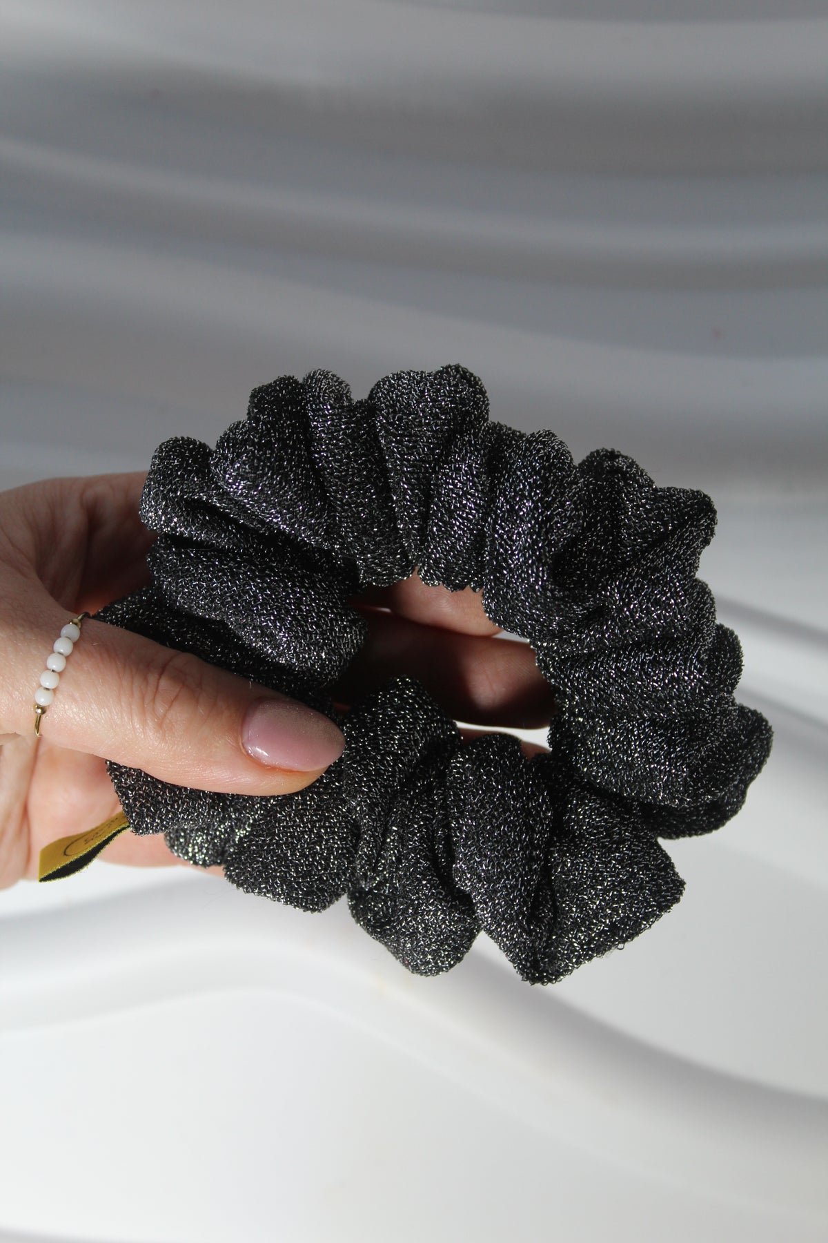 A close-up of a hand holding a black glittery scrunchie against a soft white draped fabric background.