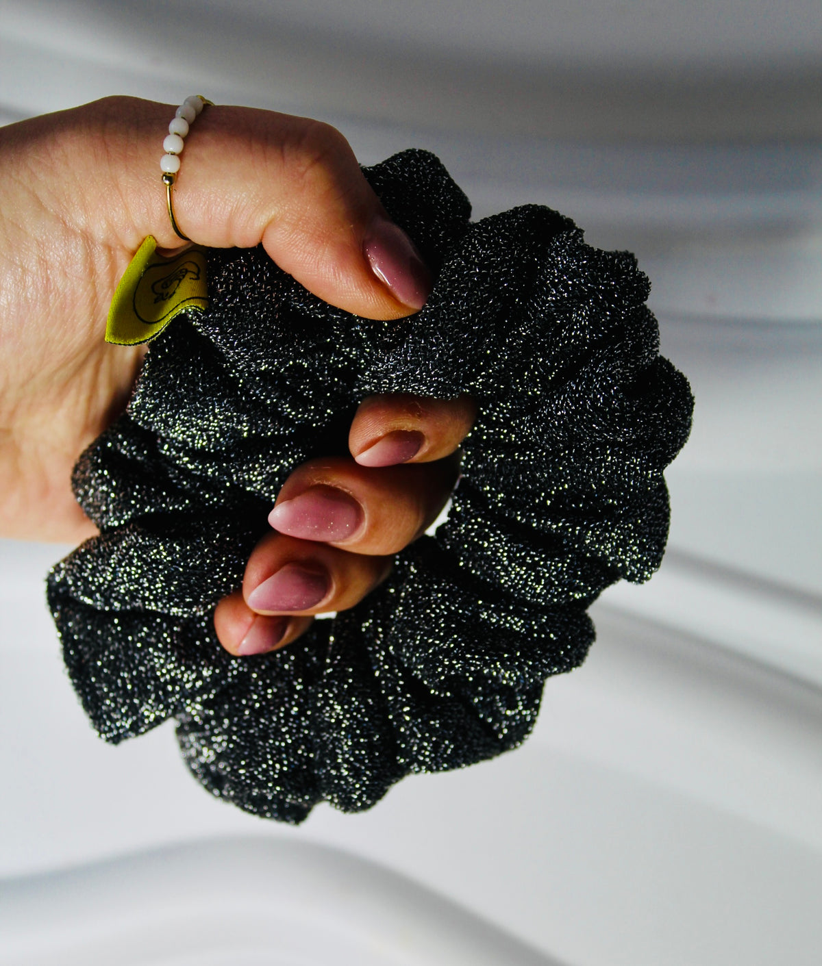 A close-up of a hand holding a black glittery scrunchie against a soft white background.