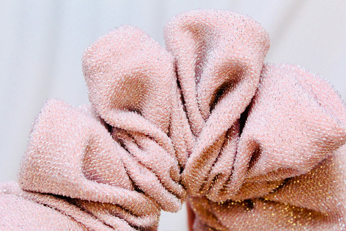 A close-up of a dusty pink scrunchie made from textured, sparkly terry cloth fabric, showing its soft folds and glittery surface.