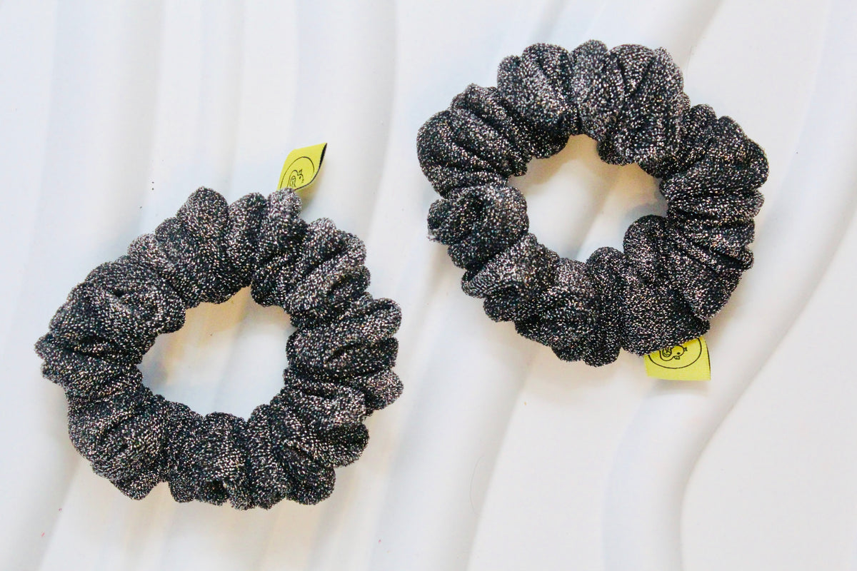 Two black glittery scrunchies with small yellow tags, displayed on a white wavy background.