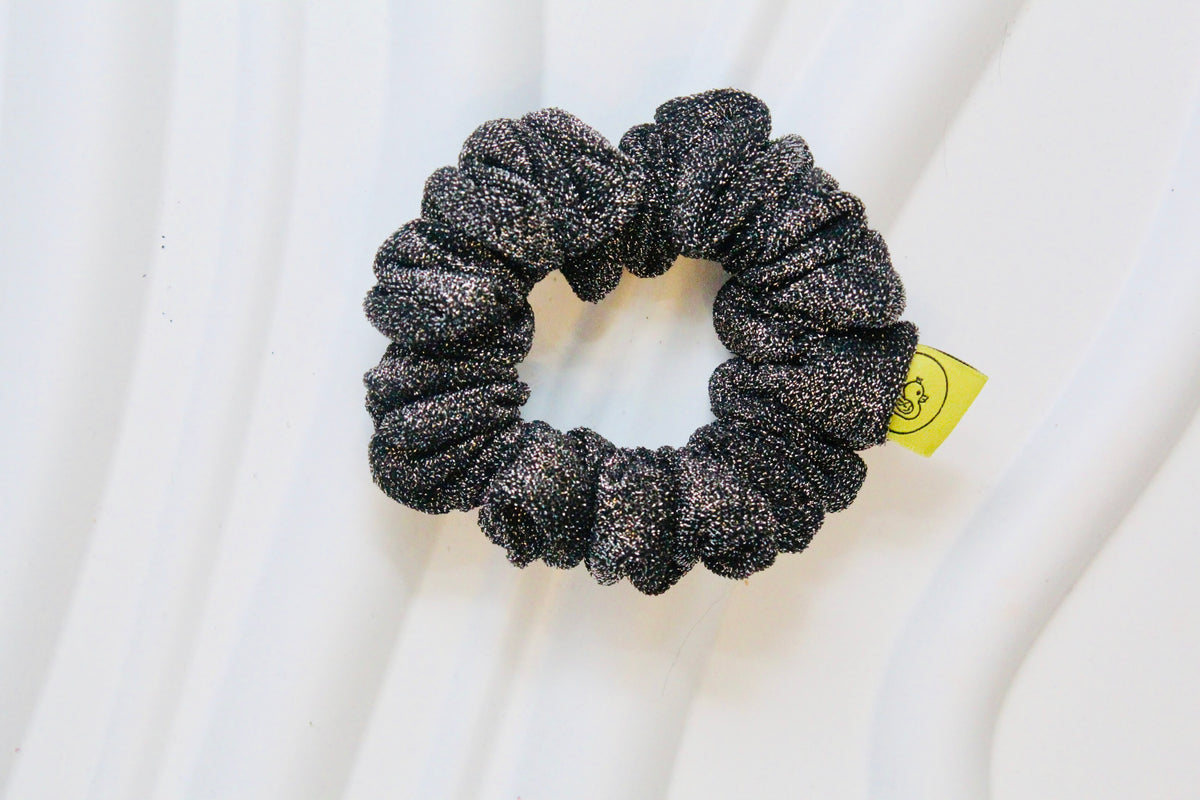 A black glittery scrunchie with a small yellow tag, displayed on a white draped fabric background.