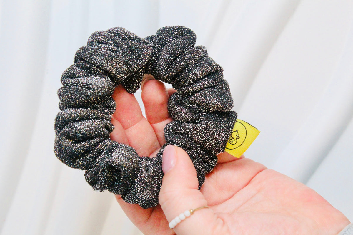 A close-up of a hand holding a black and silver glittery scrunchie with a small yellow tag attached, set against a soft white curtain background.