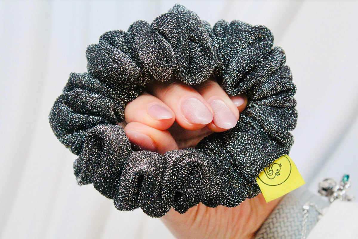 A close-up of a hand holding a dark gray glittery scrunchie with a small yellow tag attached, against a soft white background.