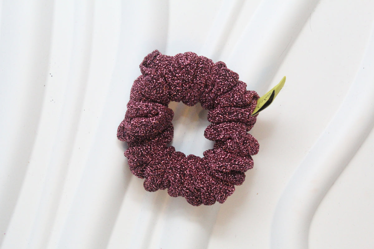 A glittery burgundy scrunchie with a small green tag, resting on a white wavy surface.
