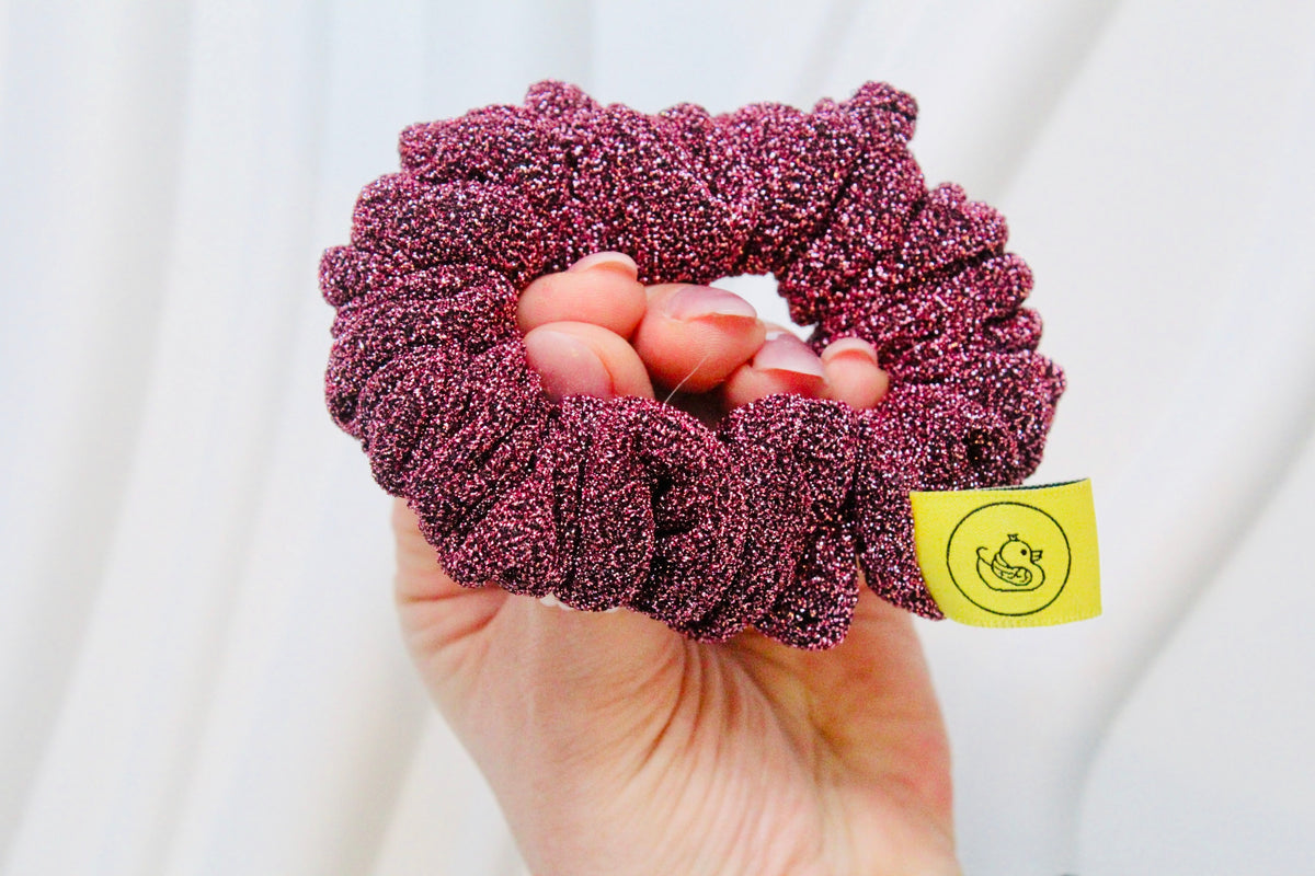 A close-up of a hand holding a sparkly, textured scrunchie in a deep berry purple color. A small yellow brand tag with a duck logo is attached to the scrunchie.