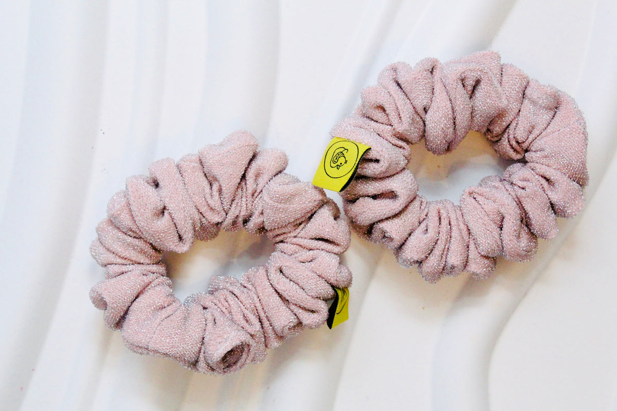 Two dusty pink textured scrunchies with small yellow tags, displayed on a white wavy background.