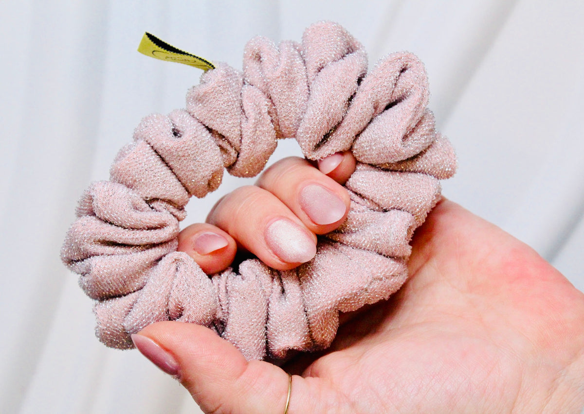 A close-up of a hand holding a dusty pink textured scrunchie with a small gold ribbon tip, set against a soft white draped fabric background.