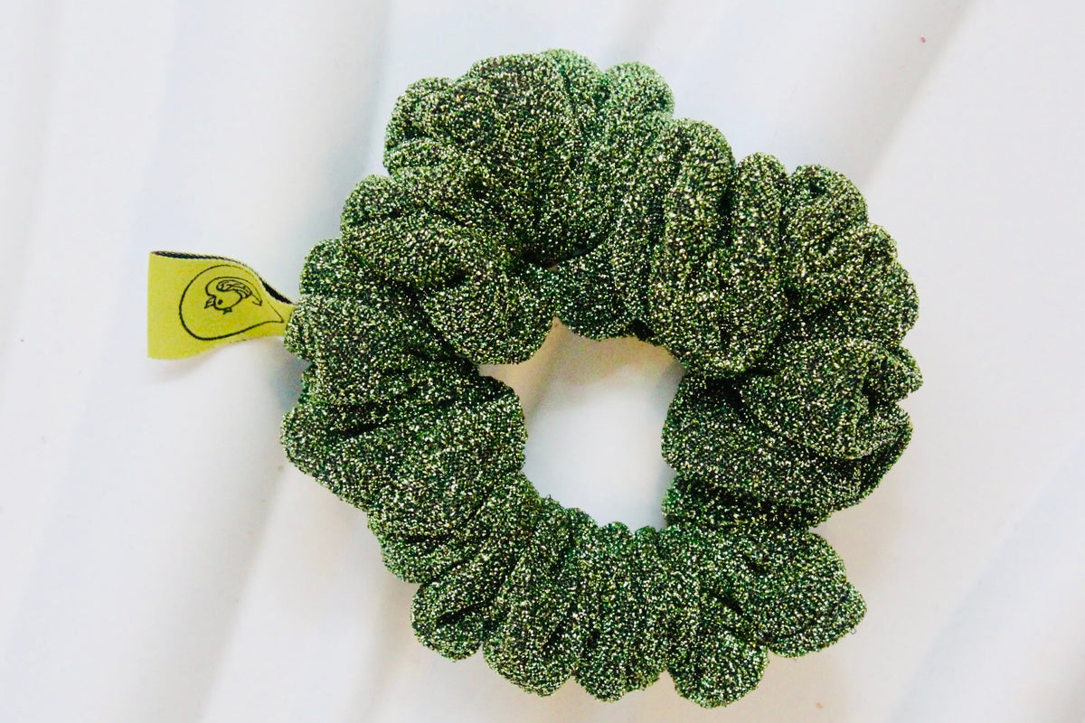 A green glittery scrunchie with a small yellow tag, photographed on a white fabric background.